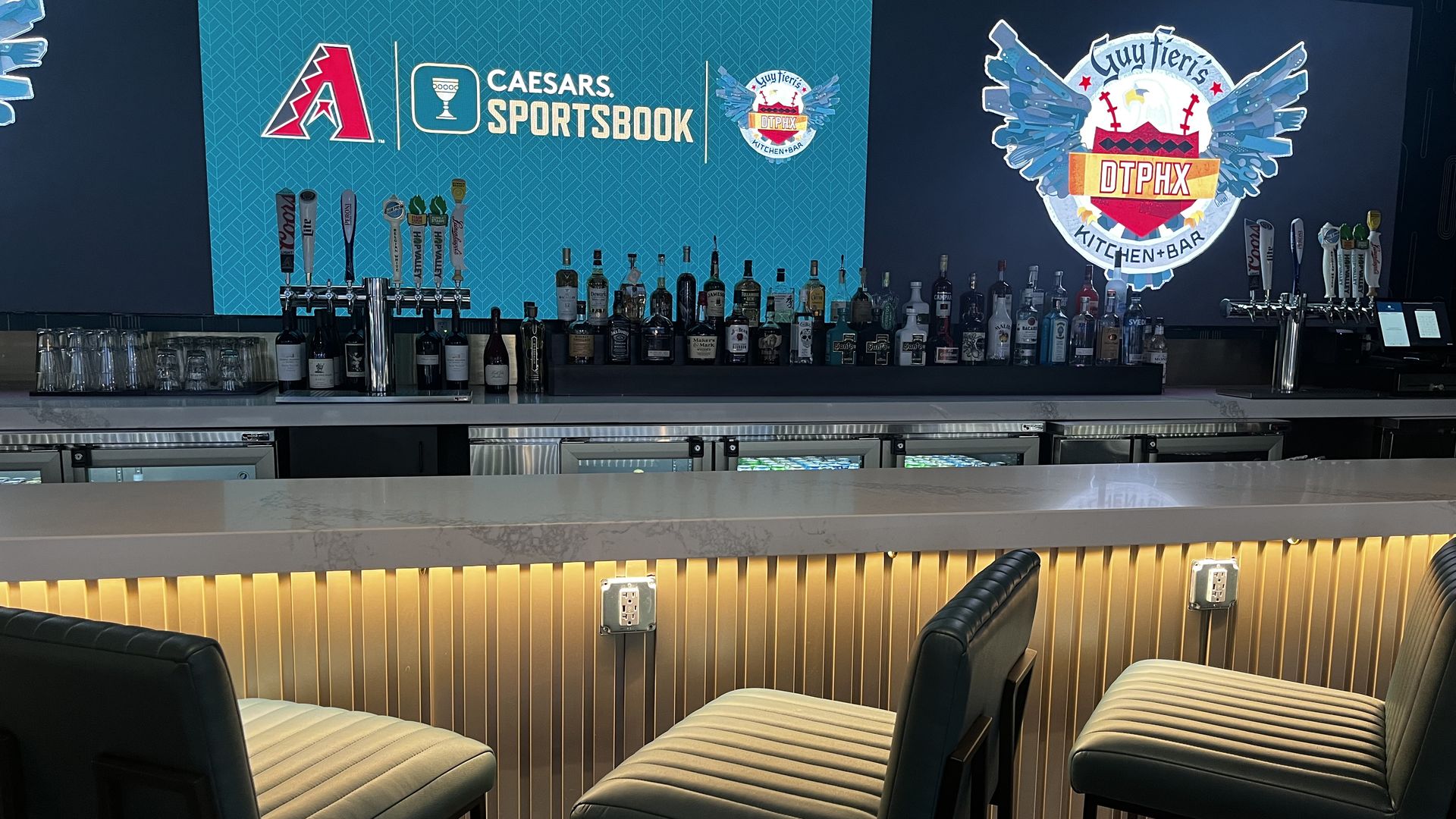 A bar and barstools in front of a screen that says "Caesars Sportsbook" with the Diamondbacks logo and a screen that says "Guy Fieri's DTPHX Kitchen Bar"