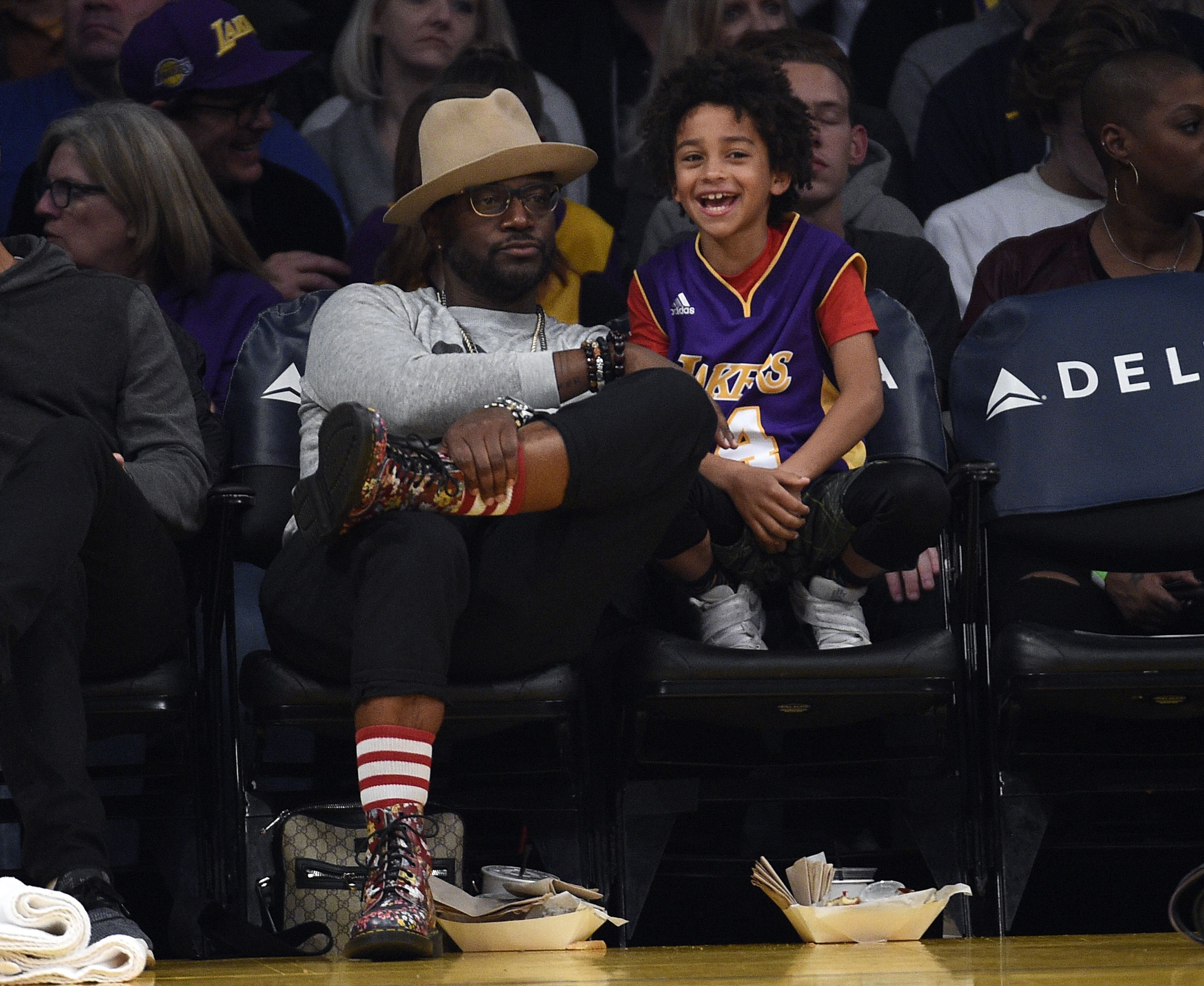Smiling child in a Lakers purple and gold jersey sits next to a man wearing a beige wide-brim hat and glasses at a basketball game; dark crowd in the background, snacks and bags on the floor nearby.