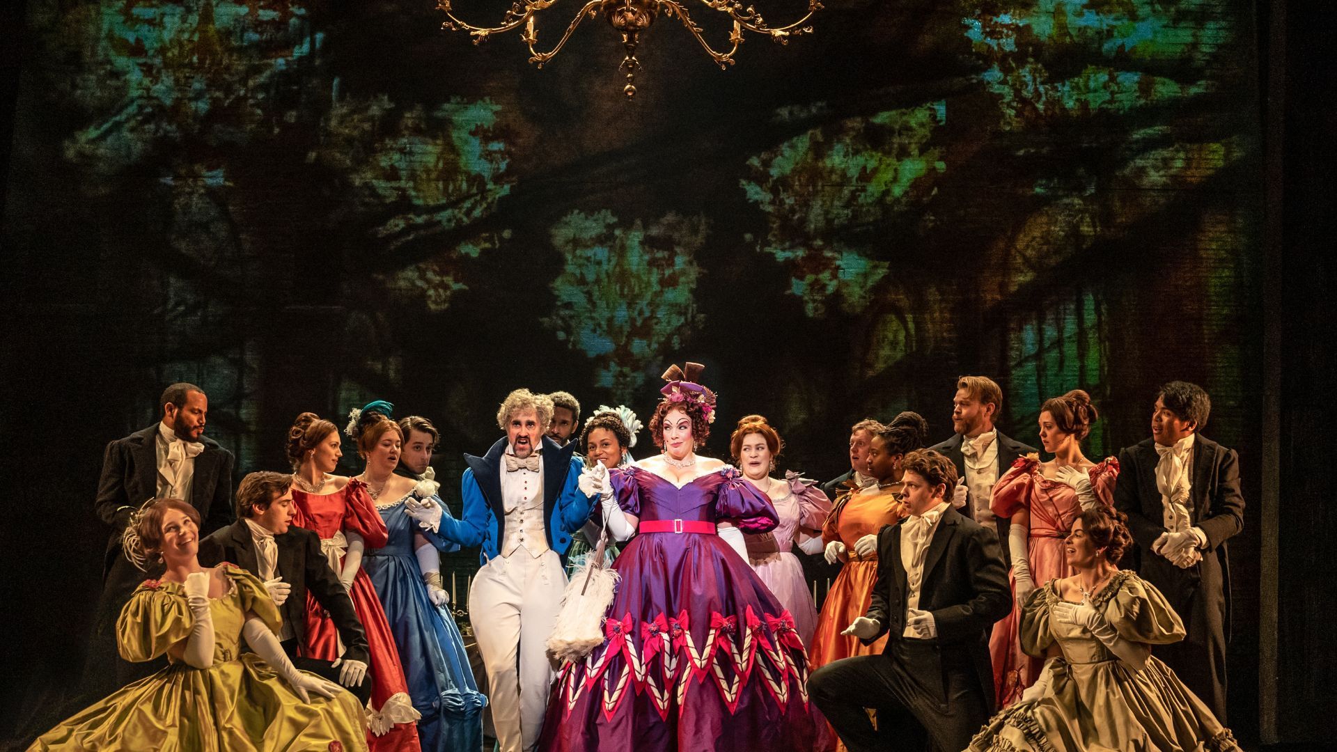 A scene from "Les Miserables" on a stage.  A large group of people are wearing formal clothing and gathered under a chandelier. 