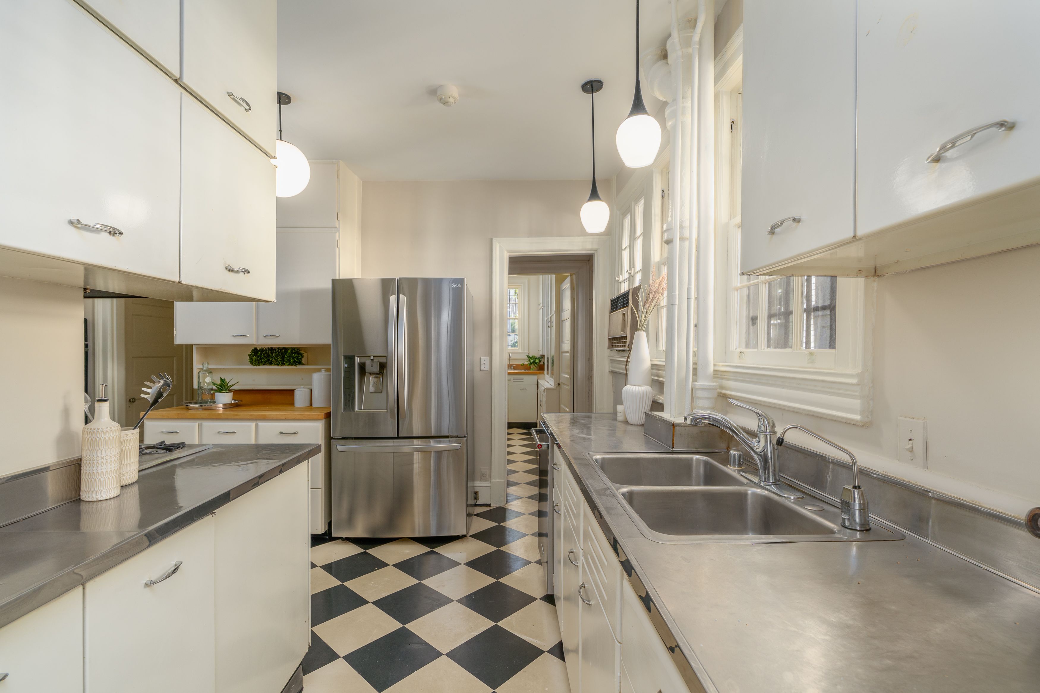 Bright galley kitchen with white cabinets, stainless steel countertops and appliances. A black-and-white checkered floor, hanging pendant lights, and a double sink along the right.