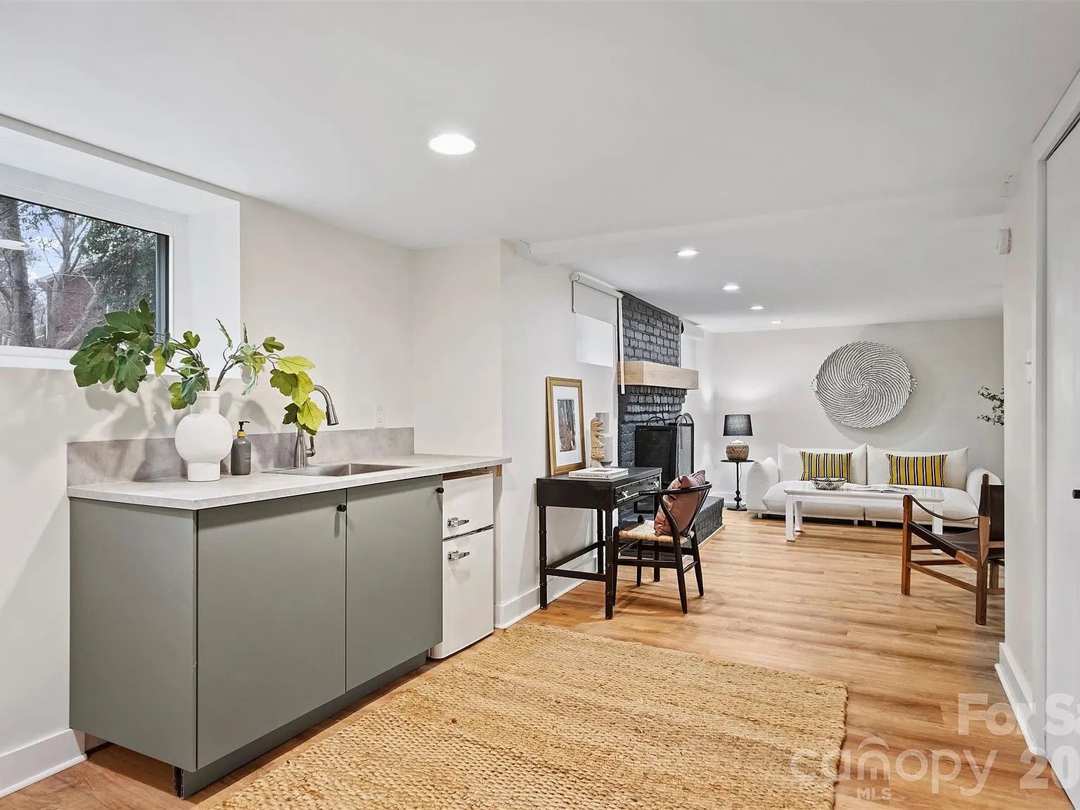 Bright modern living space with gray cabinet and sink on left, wooden floor, black brick fireplace, white sofa with yellow-striped pillows, wooden chairs, and large textured wall art.