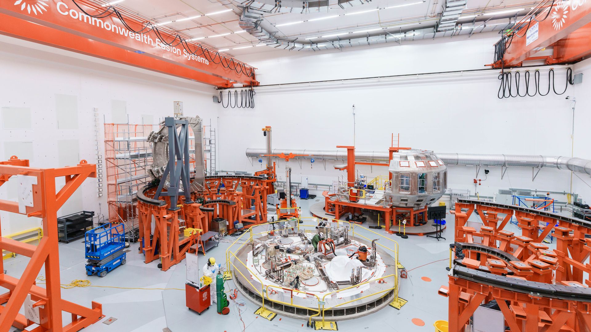 Large industrial lab with Commonwealth Fusion Systems equipment, featuring bright orange metal frames, a circular scientific apparatus, scaffolding and clean white walls with a high ceiling.