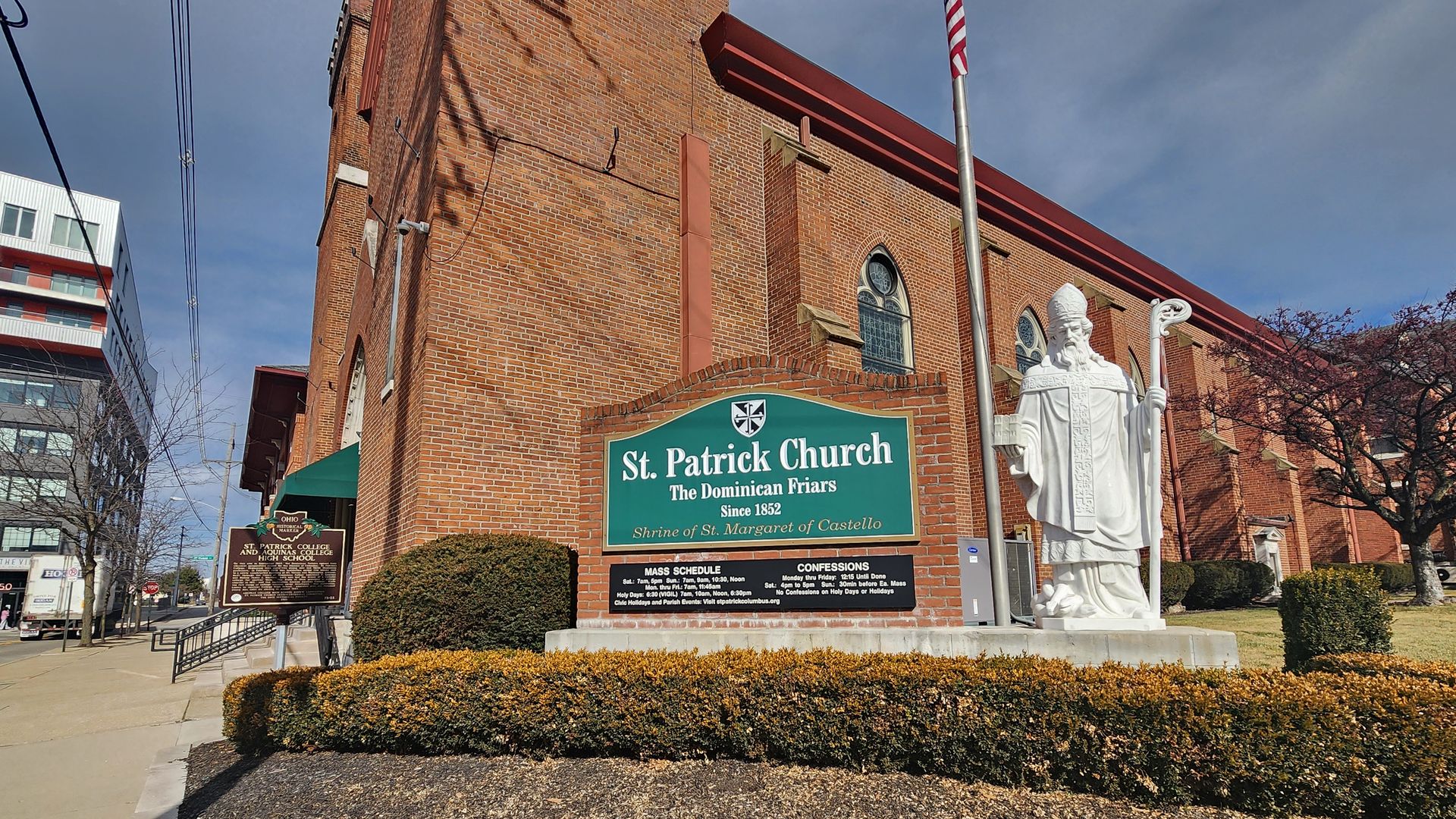 The side of St. Patrick Church with a sign, historical marker and statue of a religious figure. 