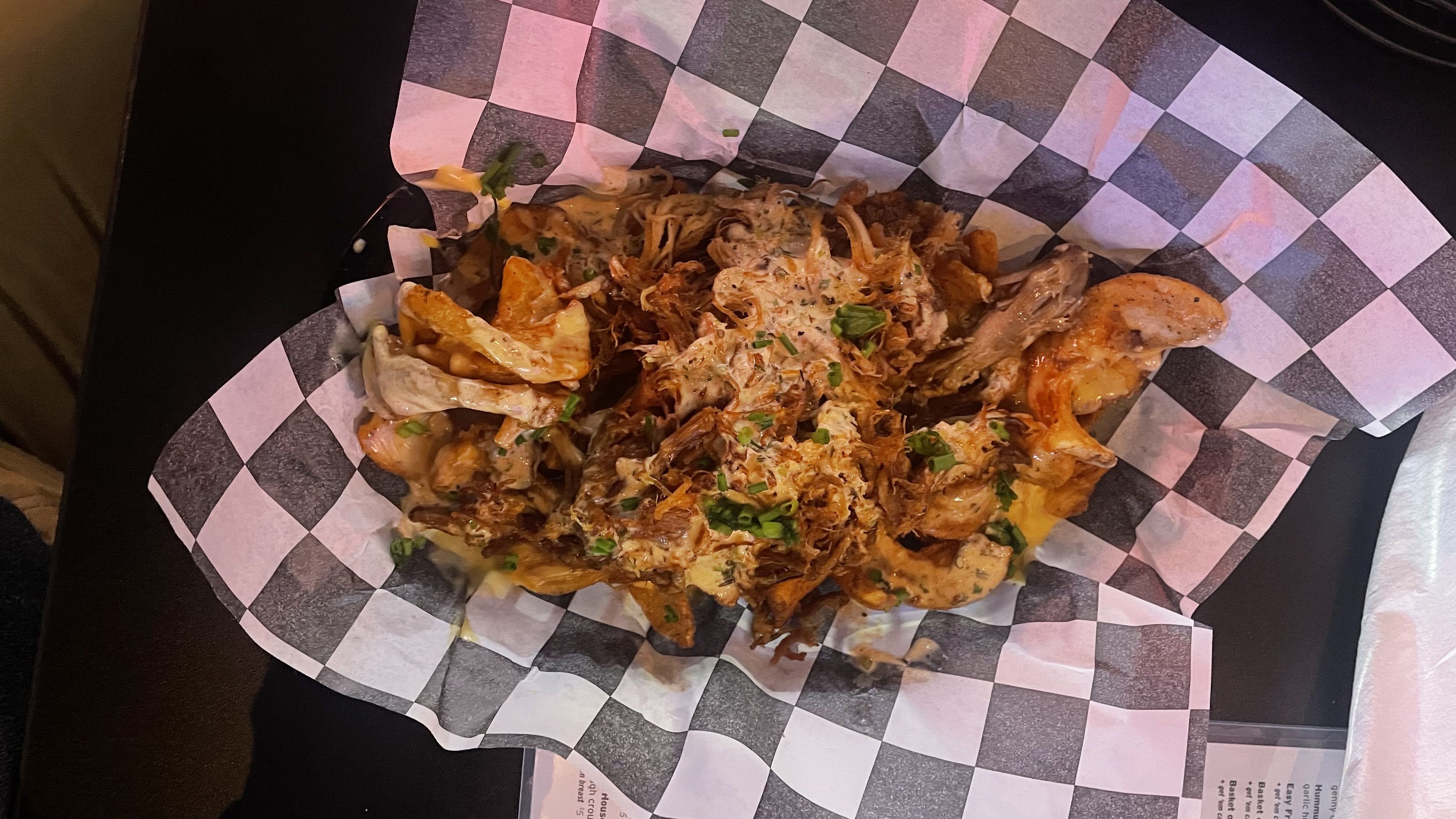 Basket of loaded sidewinder fries with melted cheese, seasoned shredded meat, creamy sauce, and garnished with chopped green onions on black and white checkered paper.