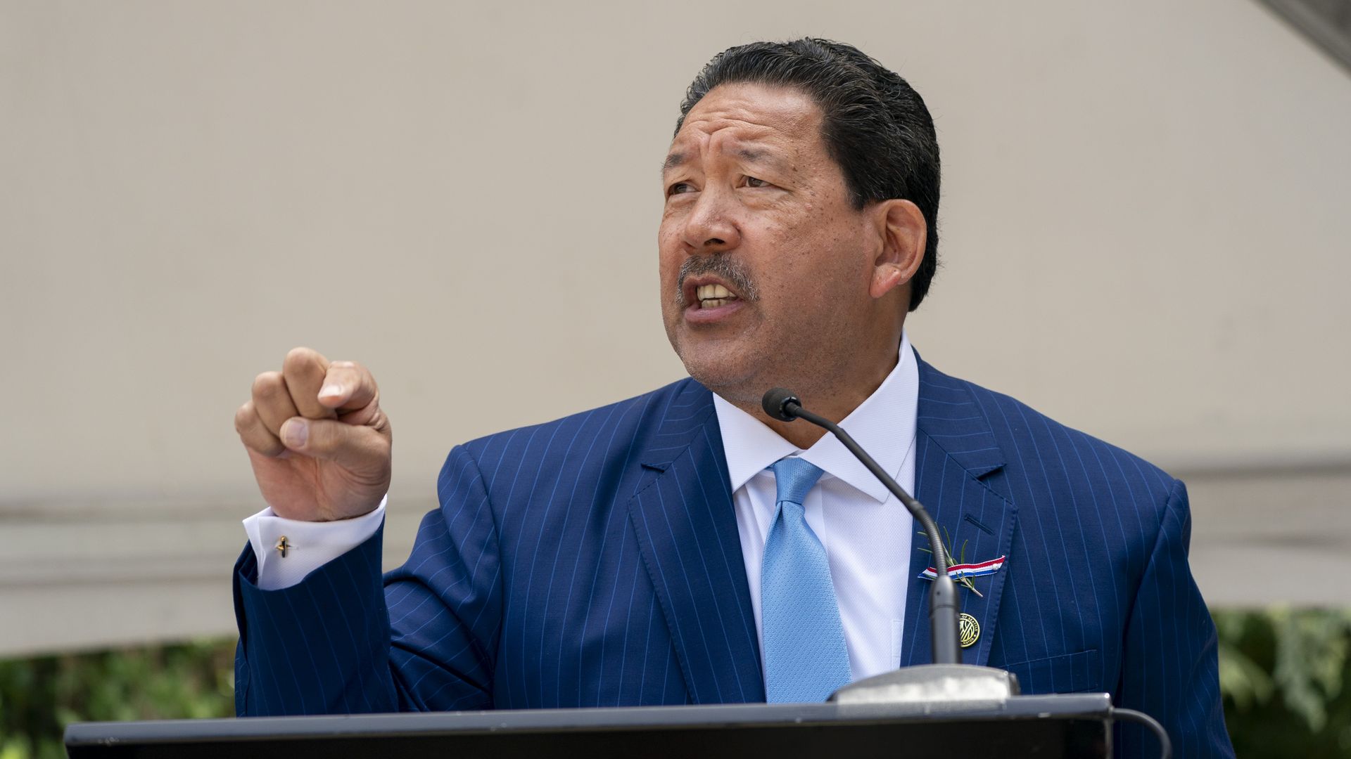 Bruce Harrell wearing a blue suit and light blue tie stands at a podium, speaking, with his finger pointed in the air.