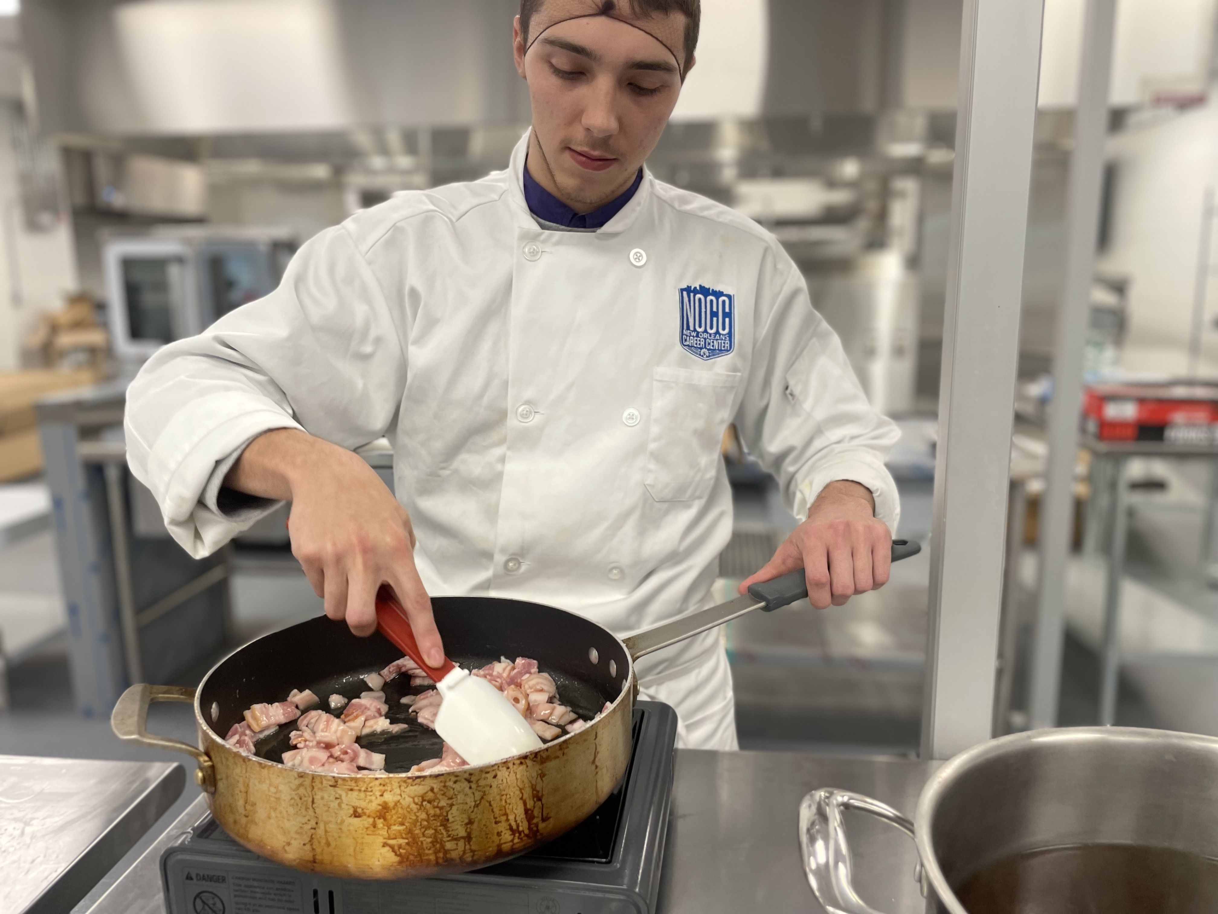 A student wearing a chef's coat uses a spatula to cook on a stove top.