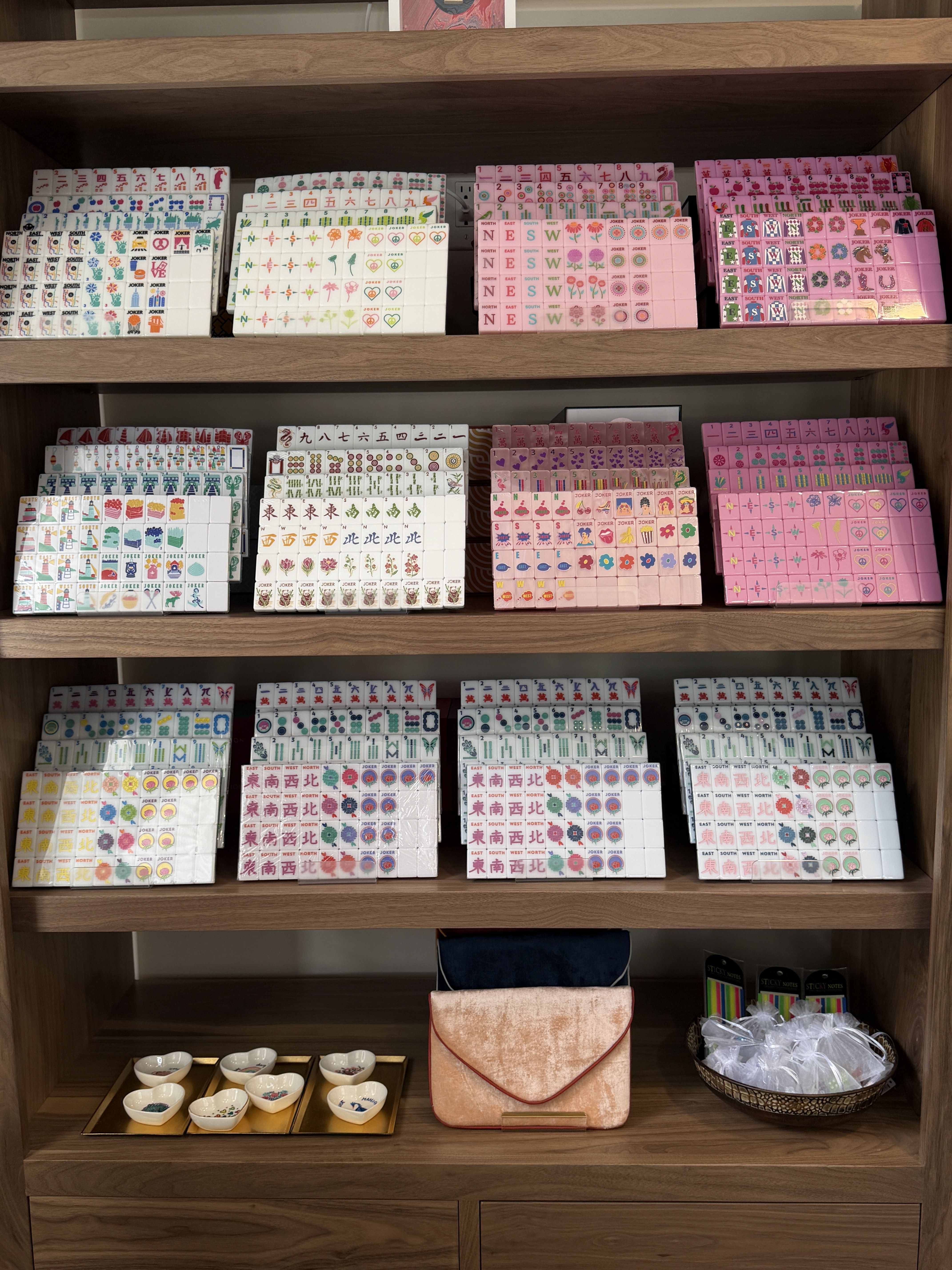 Wooden shelves stacked with colorful Mahjong tile sets in white, pink, and pastels; tiles show flowers, characters, and winds. A peach purse and small bowls sit on the lower shelf.