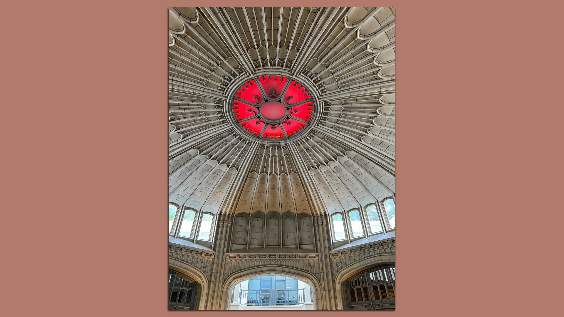 Intricate circular vaulted ceiling with radial stone ribs and a bright red circular skylight center. Arched windows surround the base, letting in daylight. Architectural detail in beige tones.