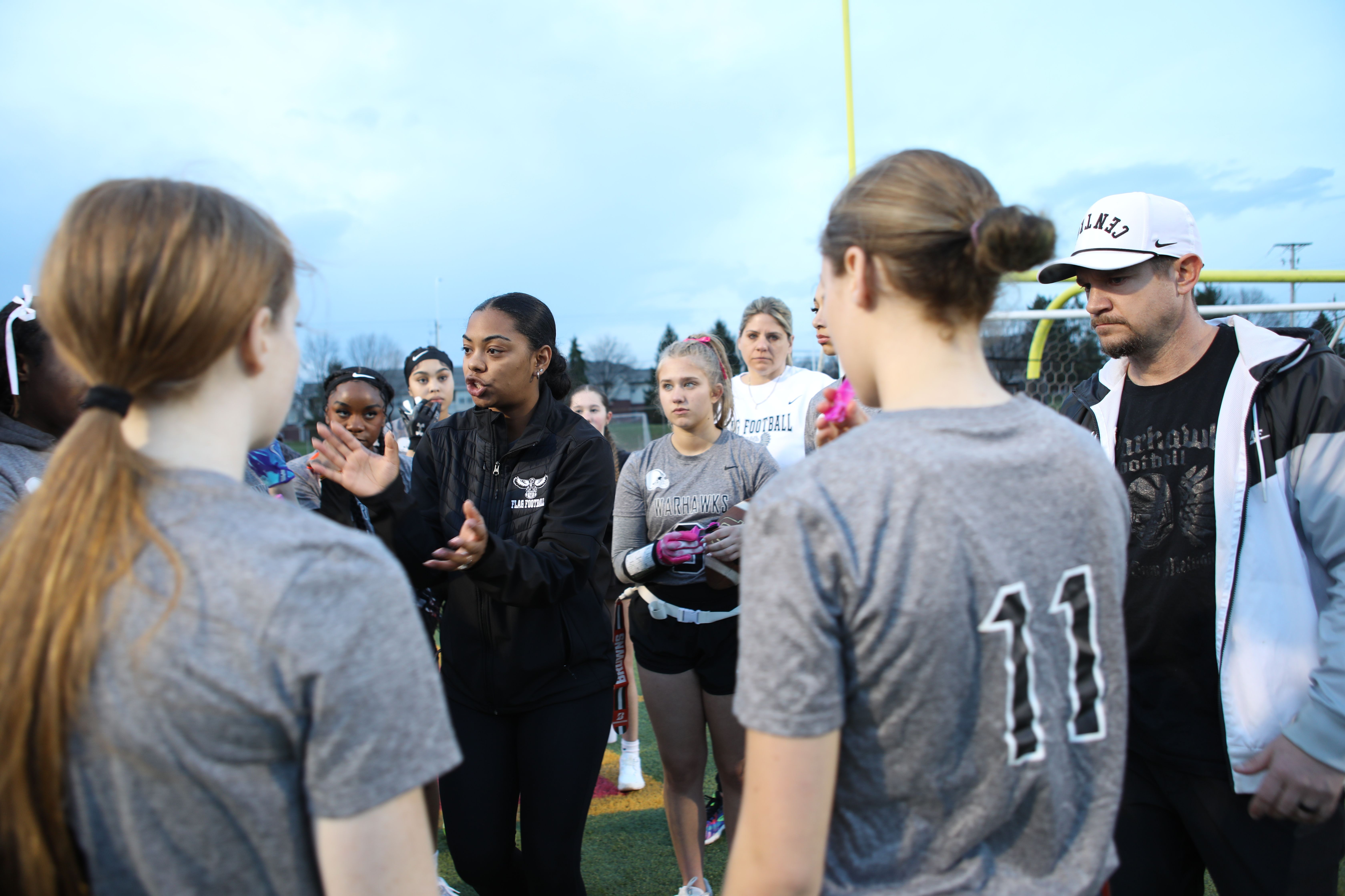 Coach in a black jacket speaks to a group of girls flag football players in gray shirts on a field, with a yellow goalpost and a blue sky behind them.