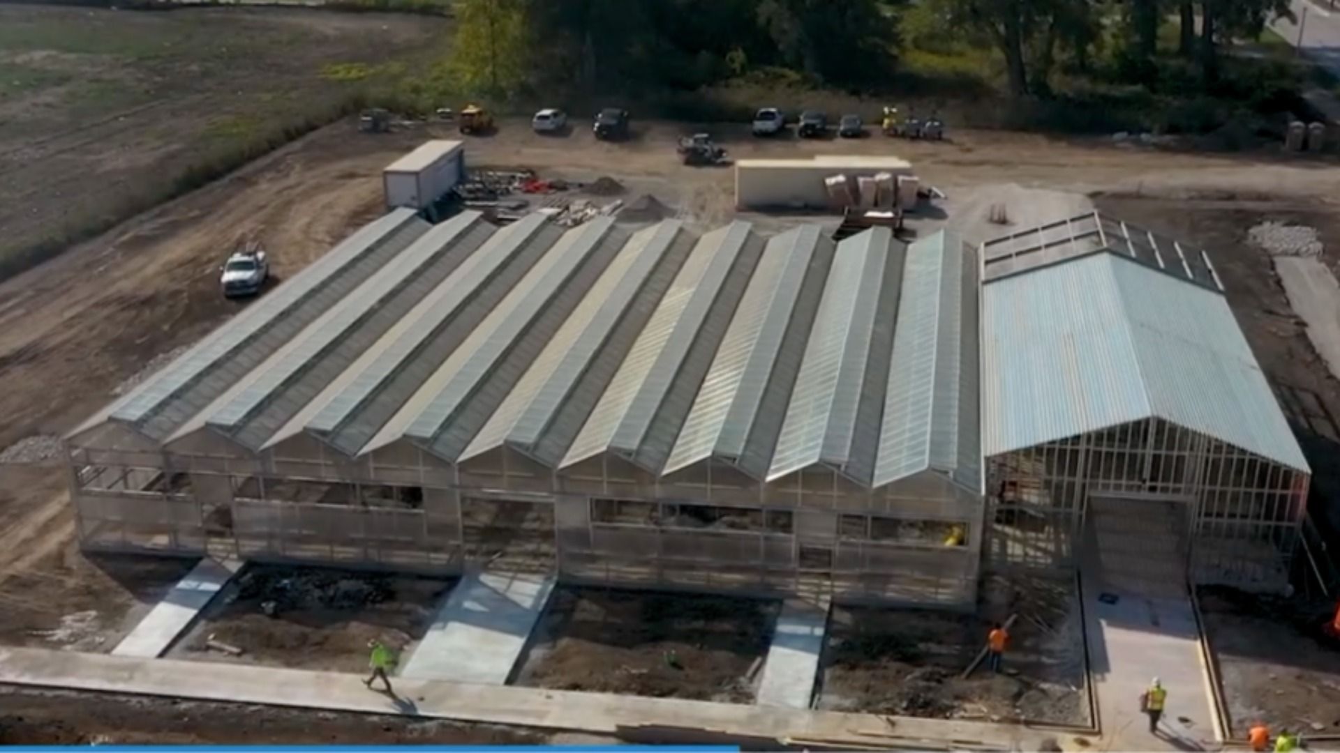 Aerial view of a large greenhouse under construction. Repeated slanted roof panels run along the length; workers in hi-vis walk a front walkway, while trucks and cars sit on a dirt lot behind.