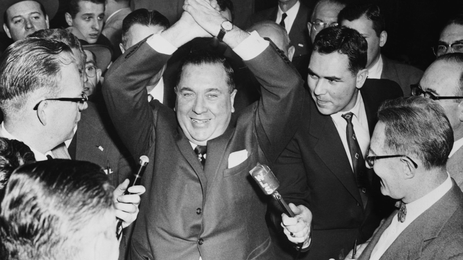 Photo of a man raising his hands in air while people with microphones surround him. 