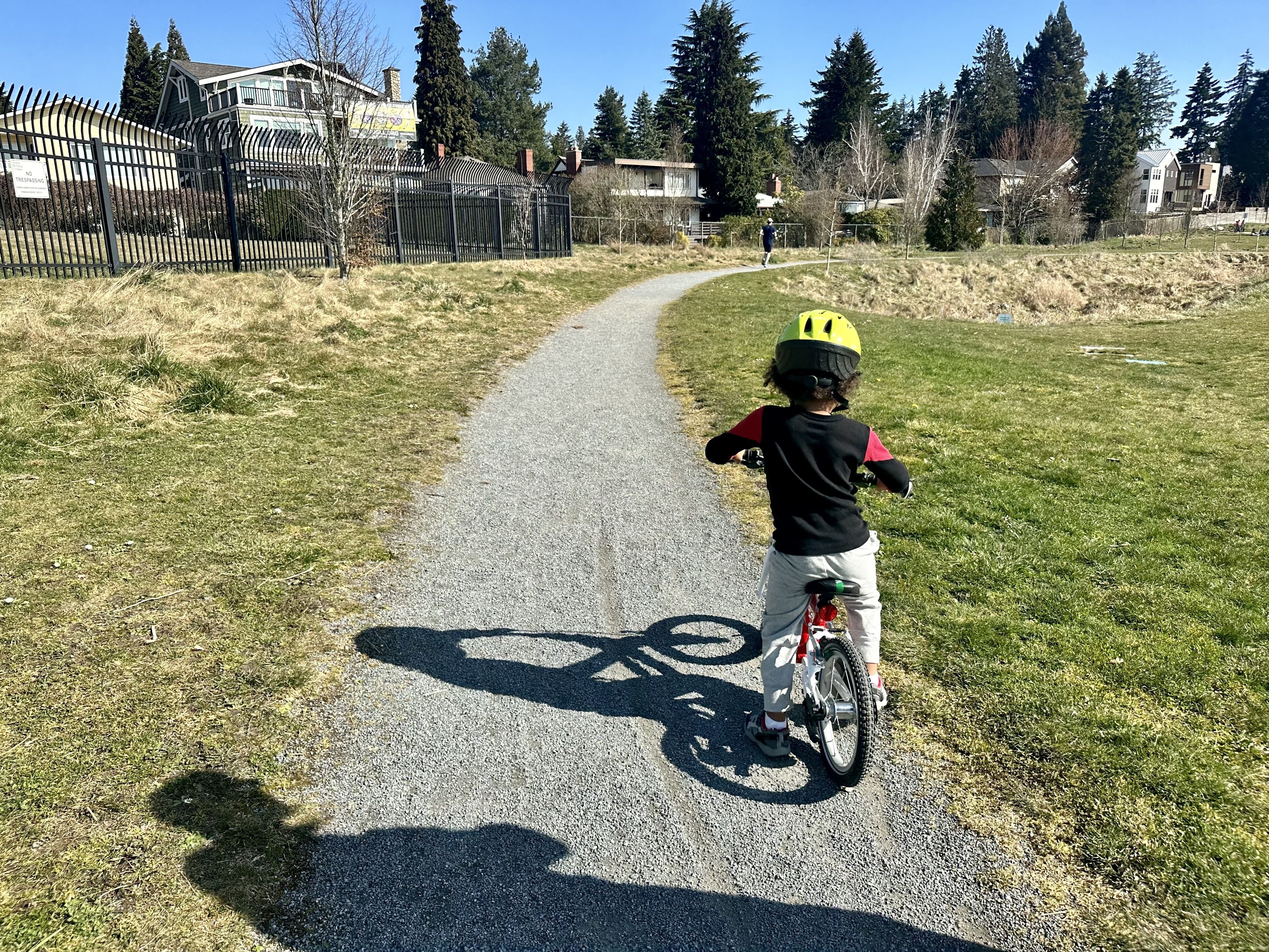 A small child in a lime green helmet straddles a bike on a gravel path, with trees and houses in the background. 