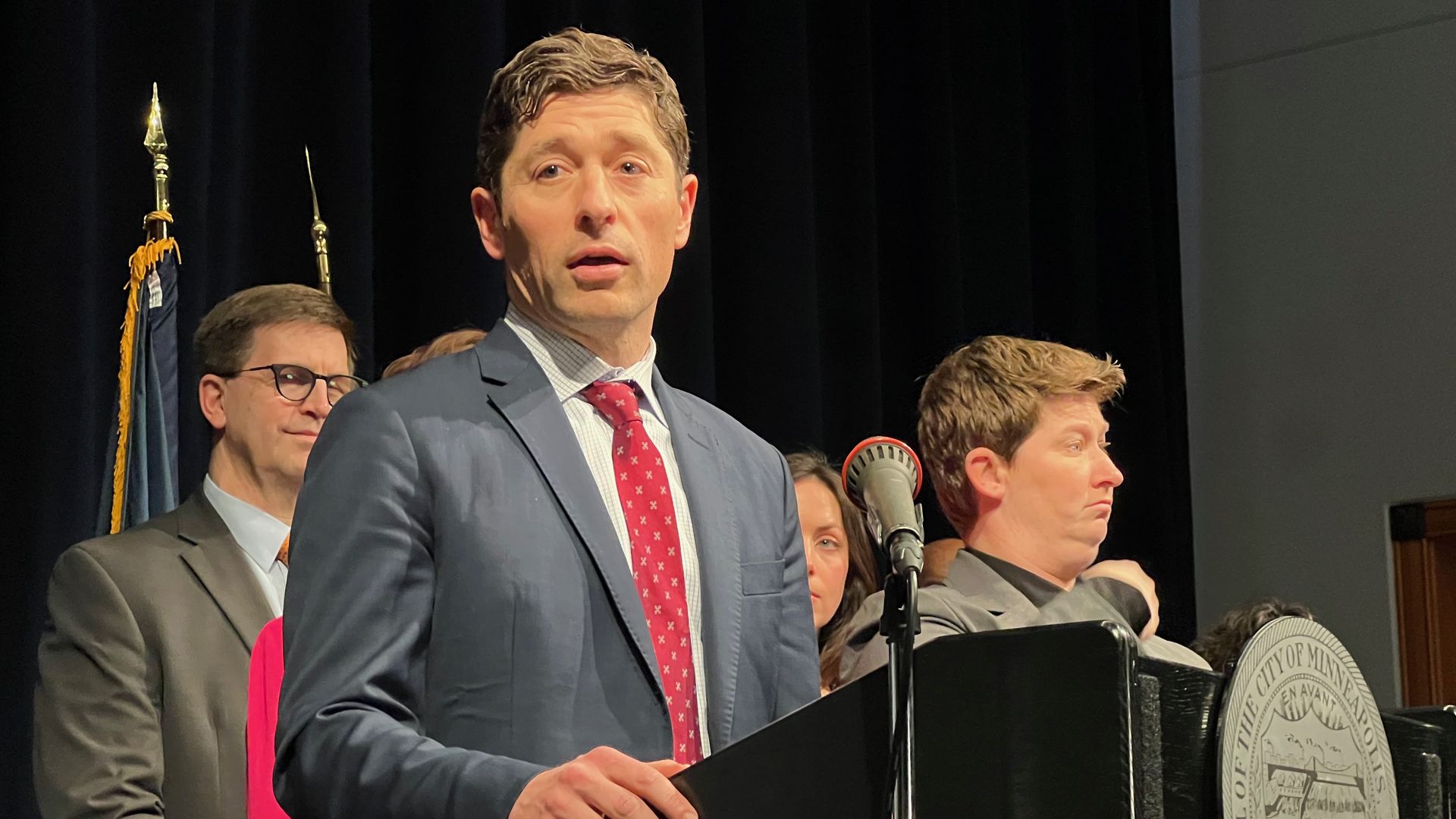 A man in a blue suit and red patterned tie speaks at a podium with a microphone during a formal event. Dark stage curtains behind him, with a few people and a city seal visible on the podium.
