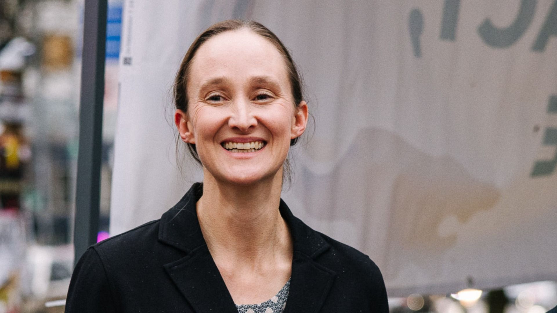 Smiling woman with light skin and pulled-back hair wearing a black coat and patterned top, standing outdoors in a city setting with blurred background.