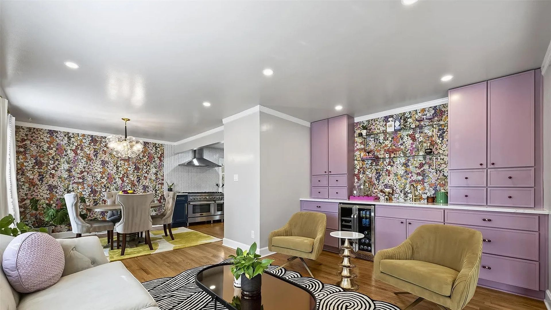 Bright living area with white sofa, lavender round pillow, black-and-white patterned rug, mustard yellow chairs, lavender cabinets with floral backsplash, and a dining area with floral wallpaper.