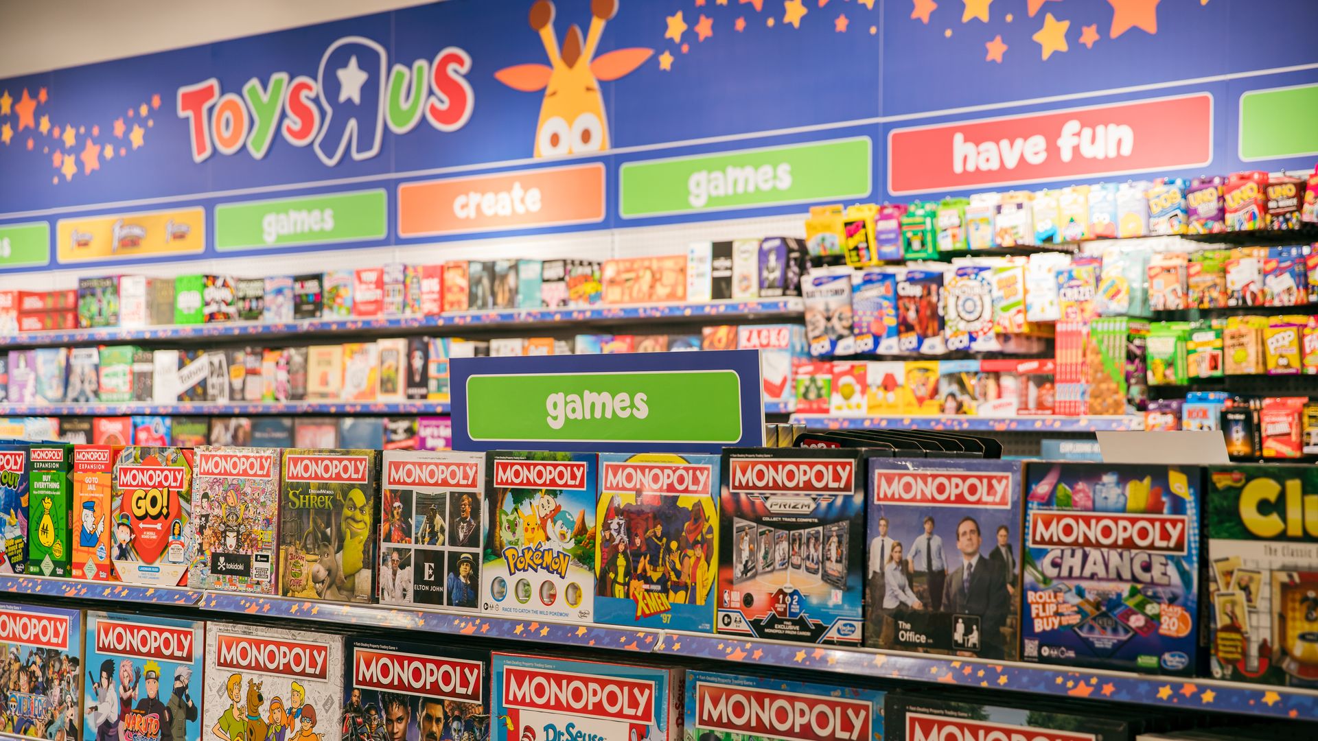 Shelves full of colorful Monopoly board game editions at Toys"R"Us, under green and orange signs saying "games" and "create" with the Toys"R"Us giraffe mascot on a blue wall.