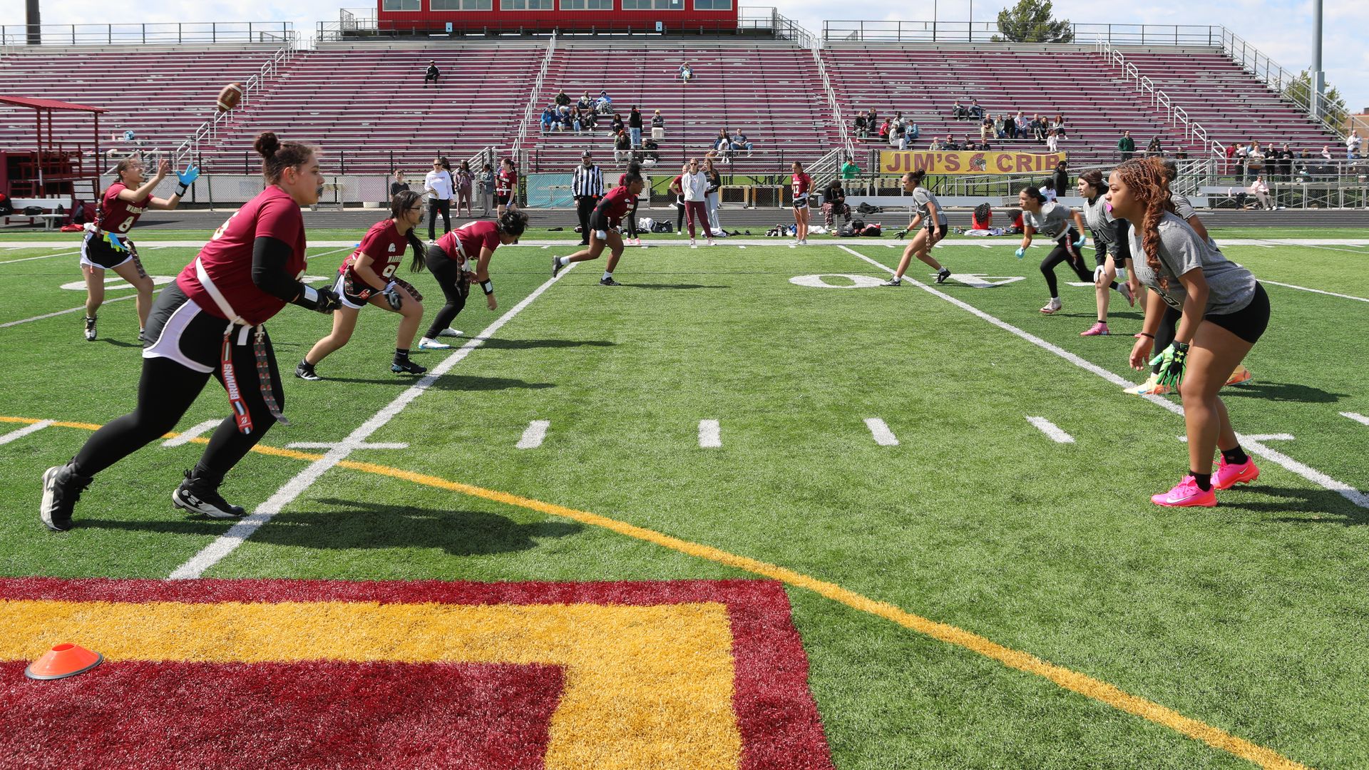 Sunlit football field with girls flag football players in maroon jerseys sprinting on green turf and players in gray jerseys defending; red bleachers and a press box rise behind.