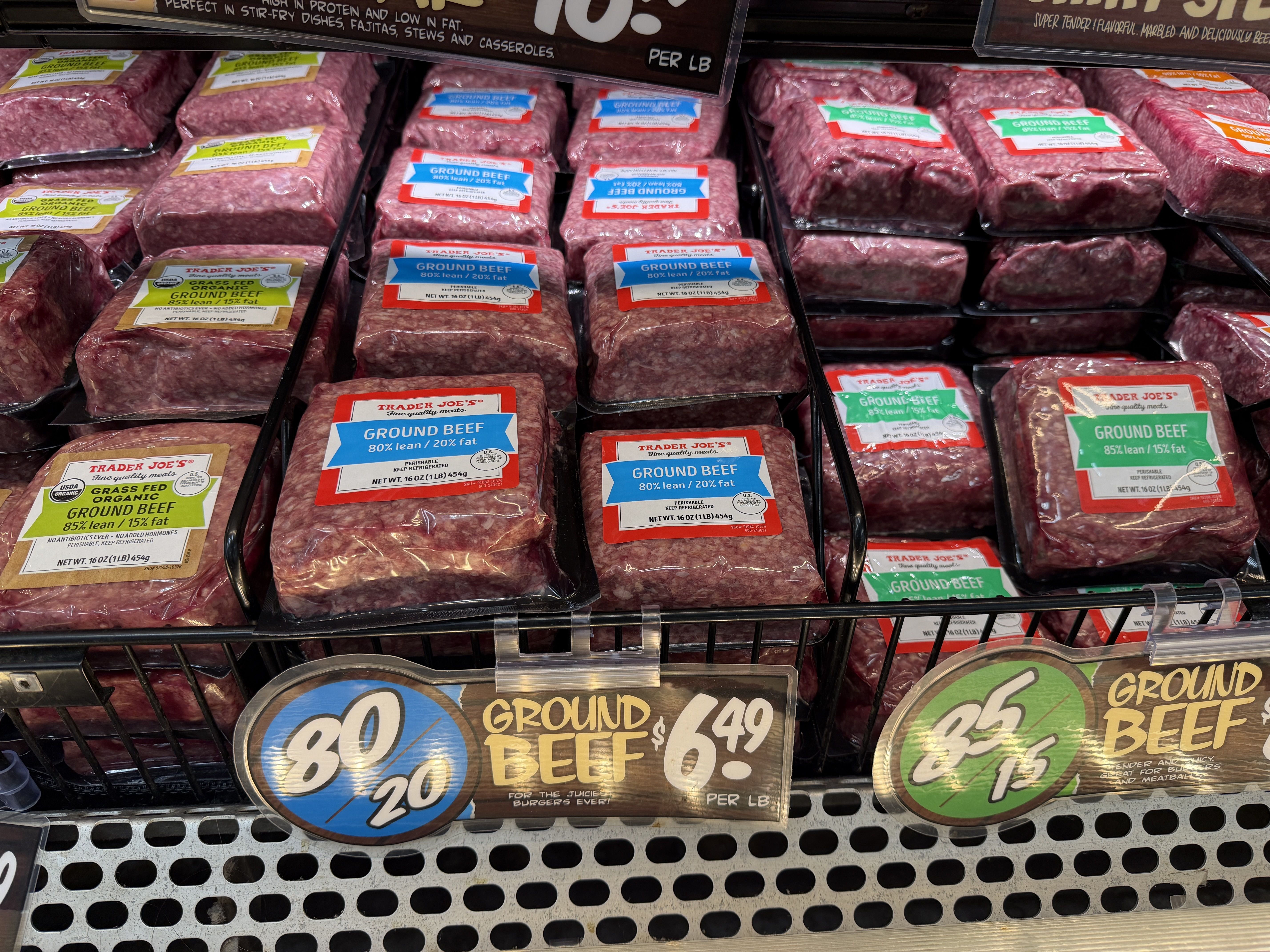 Meat case filled with ground beef packages in plastic wrap. Labels are red, blue, and green reading "Ground Beef" and "Trader Joe's"; large round price signs sit below.