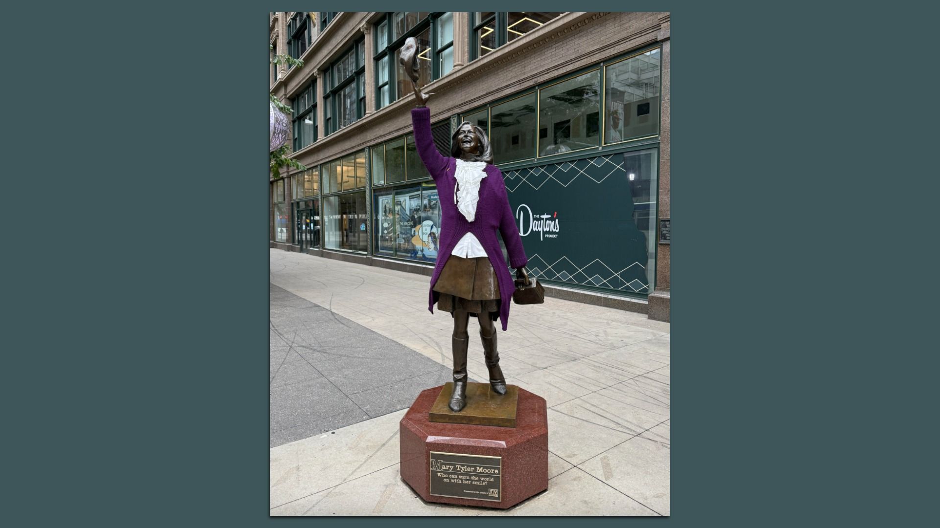 Bronze statue of Mary Tyler Moore in a purple coat waving her hat, standing on a red pedestal with an inscription, outside a building with large windows and a sign reading "The Dayton's Project".