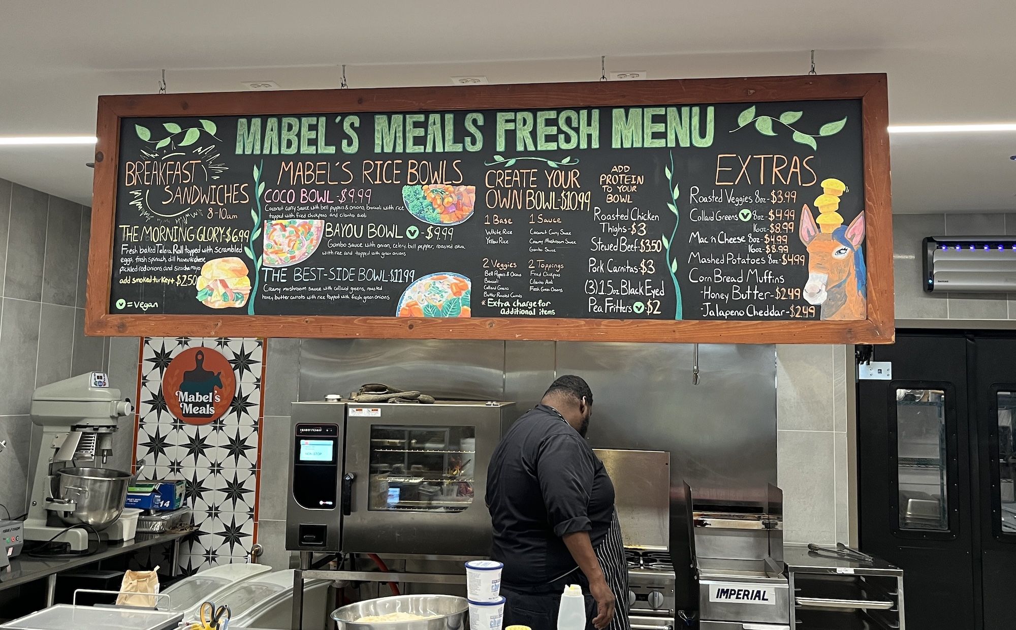 Chalkboard menu reading MABEL'S MEALS FRESH MENU hangs above a kitchen. A chef in a black shirt works at stainless appliances; a mixer and oven are visible in the scene.