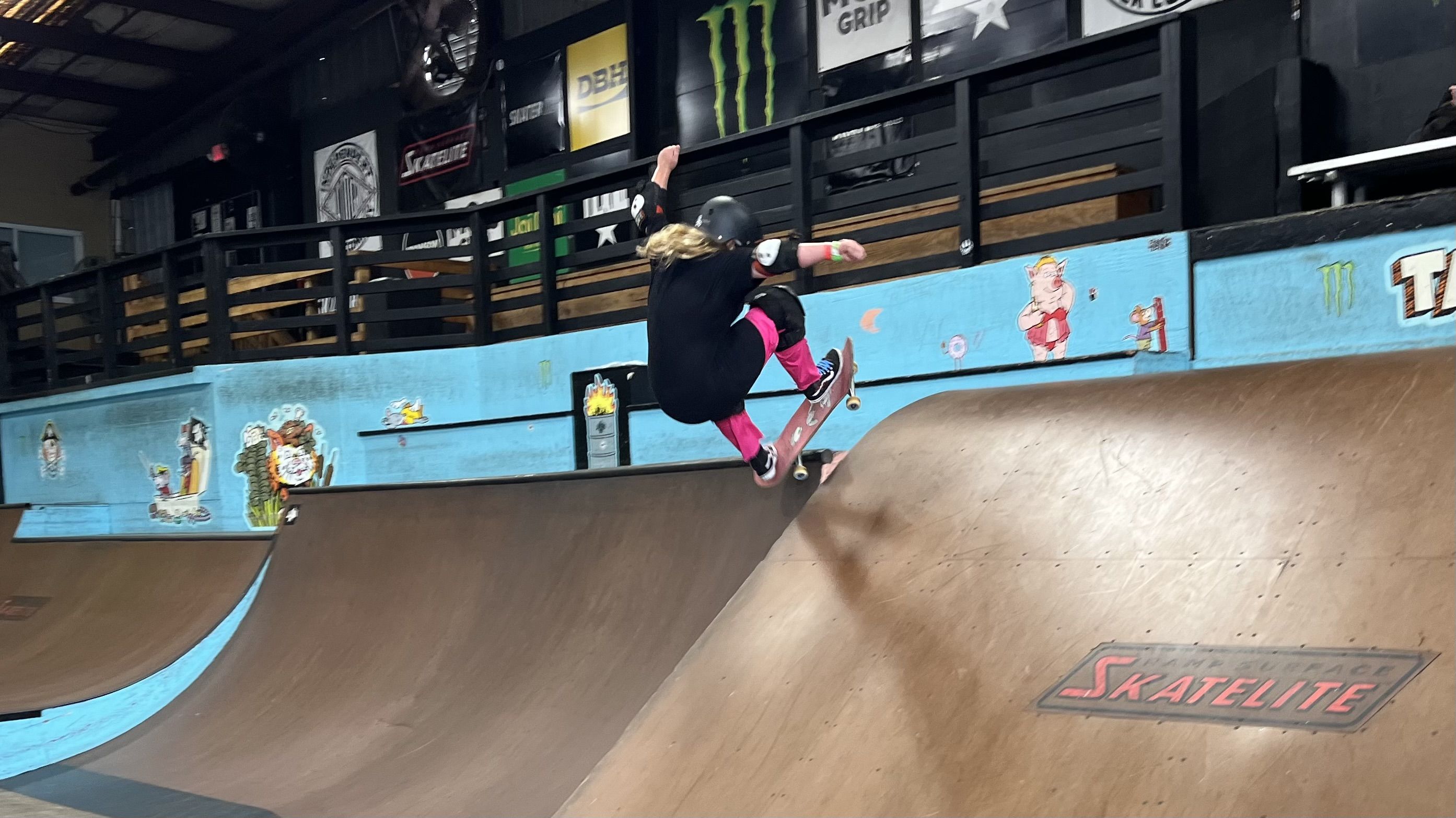 A girl in pink pants and black skirt catching air on a skateboard.
