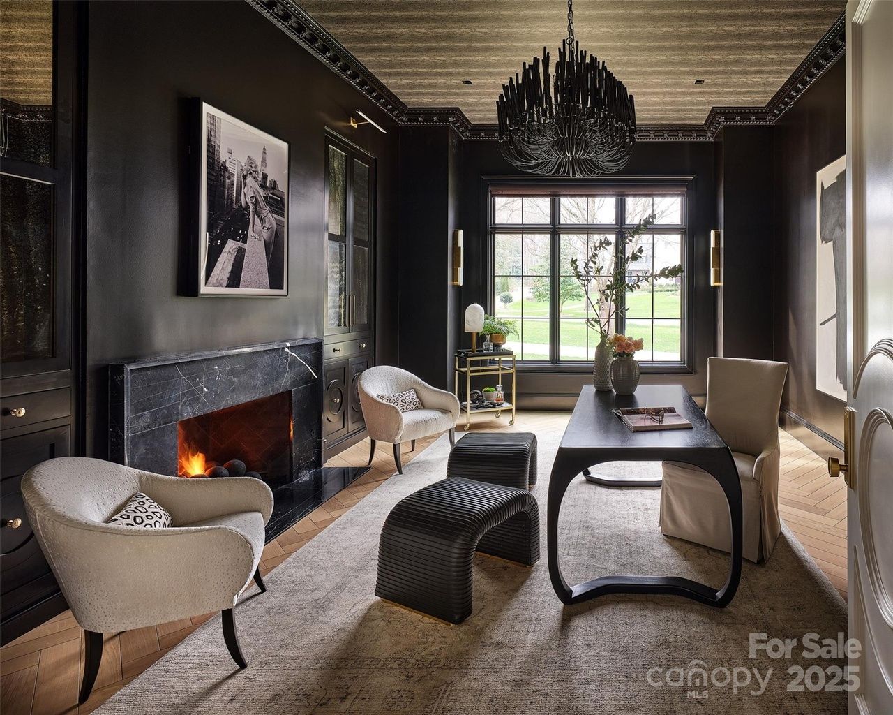 Elegant dark room with black walls, a black marble fireplace with fire, beige armchairs, modern black coffee table, large window, chandelier, and decor with plants and artwork.