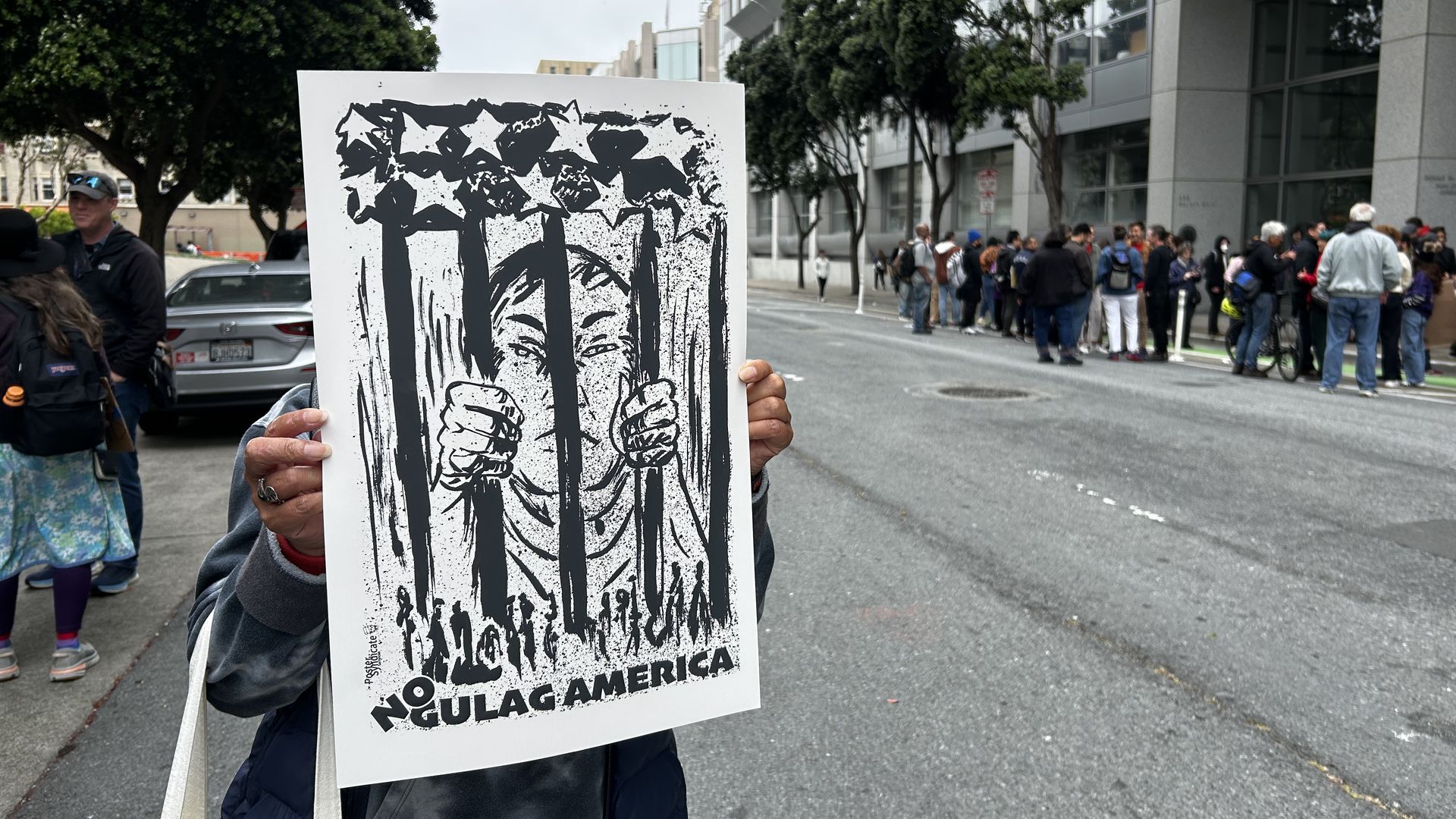 Photo of a person holding up a sign with a picture of someone behind bars and the phrase "No Gulag America." The person is standing on a street with people crowded in the background.