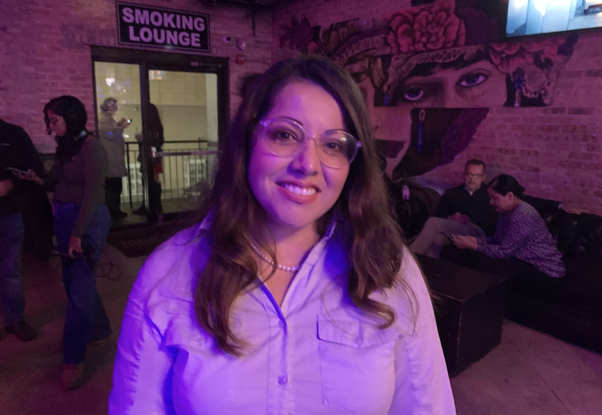 Woman in white button down shirt and glasses poses at a bar with a sign over a door reading Smoking Lounge.