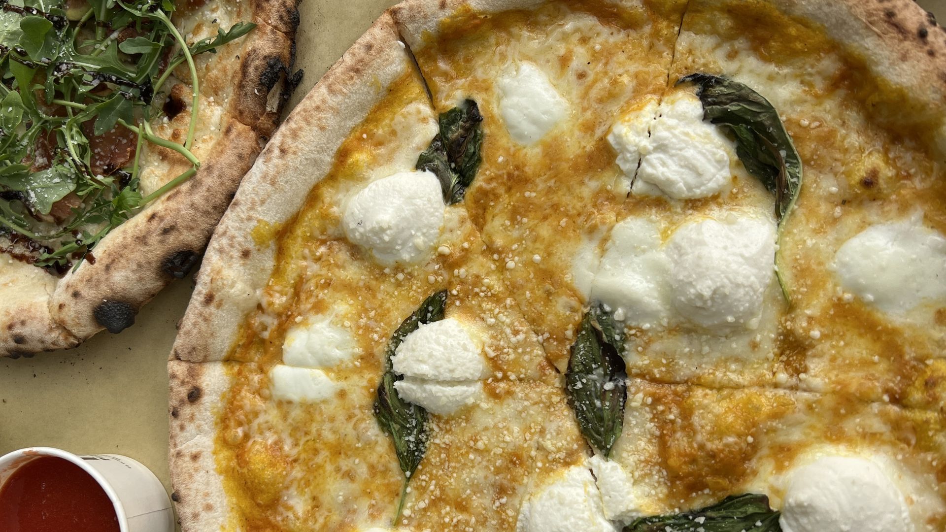 Close-up of a Margherita pizza with melted cheese, dollops of ricotta, and basil leaves on a golden crust; part of another pizza with arugula and a cup of red sauce visible on the side.