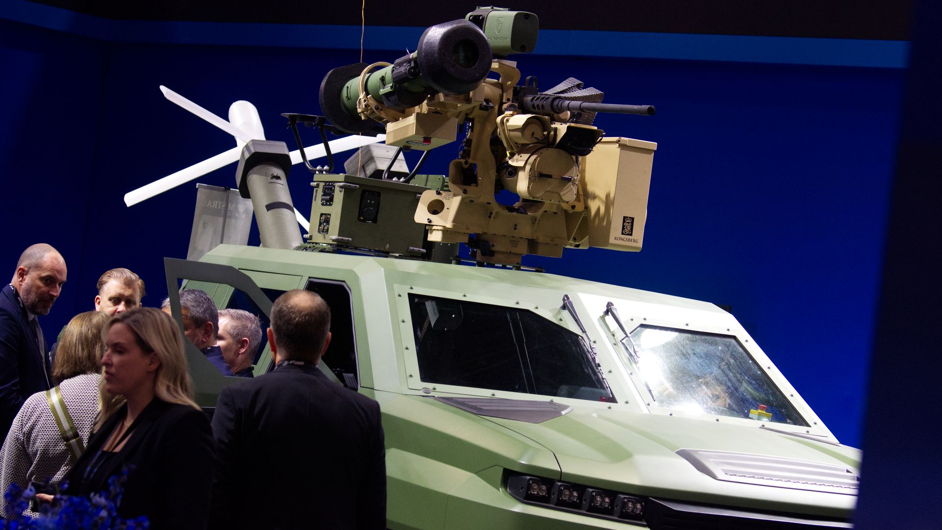 A pickup truck modified for war. It's green. The background is blue. People walk past it.