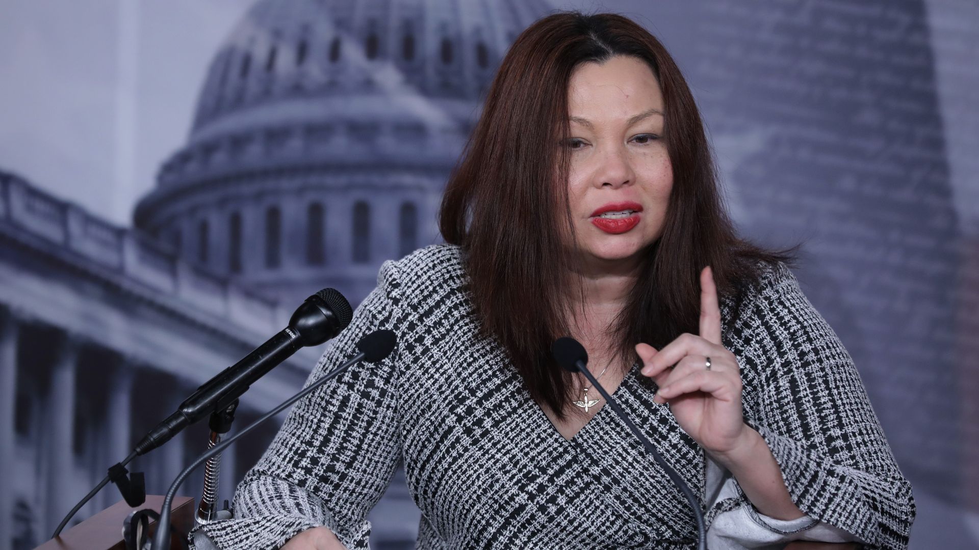 A close-up of Sen. Tammy Duckworth addressing reporters from a podium, waving her left index finger 