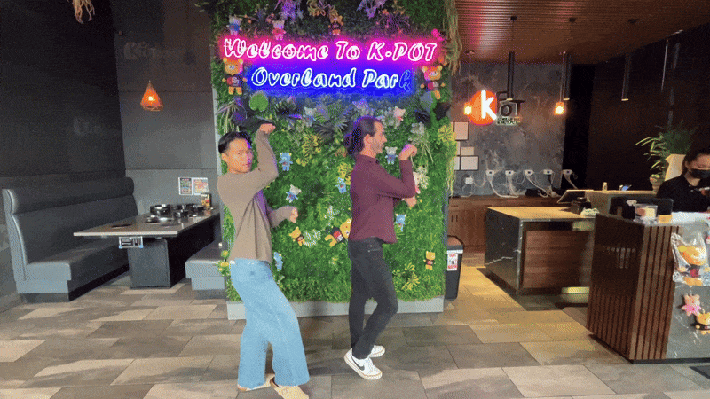 Reporter Travis and K-pop dancer Jaye Xaymongkhonh do synchronized dance moves in front of a lush green wall at KPOT in Overland Park.