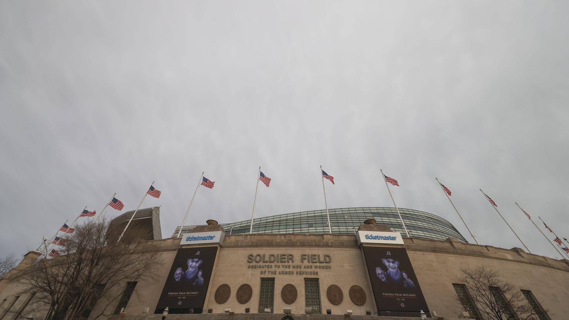 Exterior of Soldier Field stadium under a cloudy sky, featuring rows of American flags, large banners honoring Virginia Halas McCaskey, and a dedication to armed services on the facade.