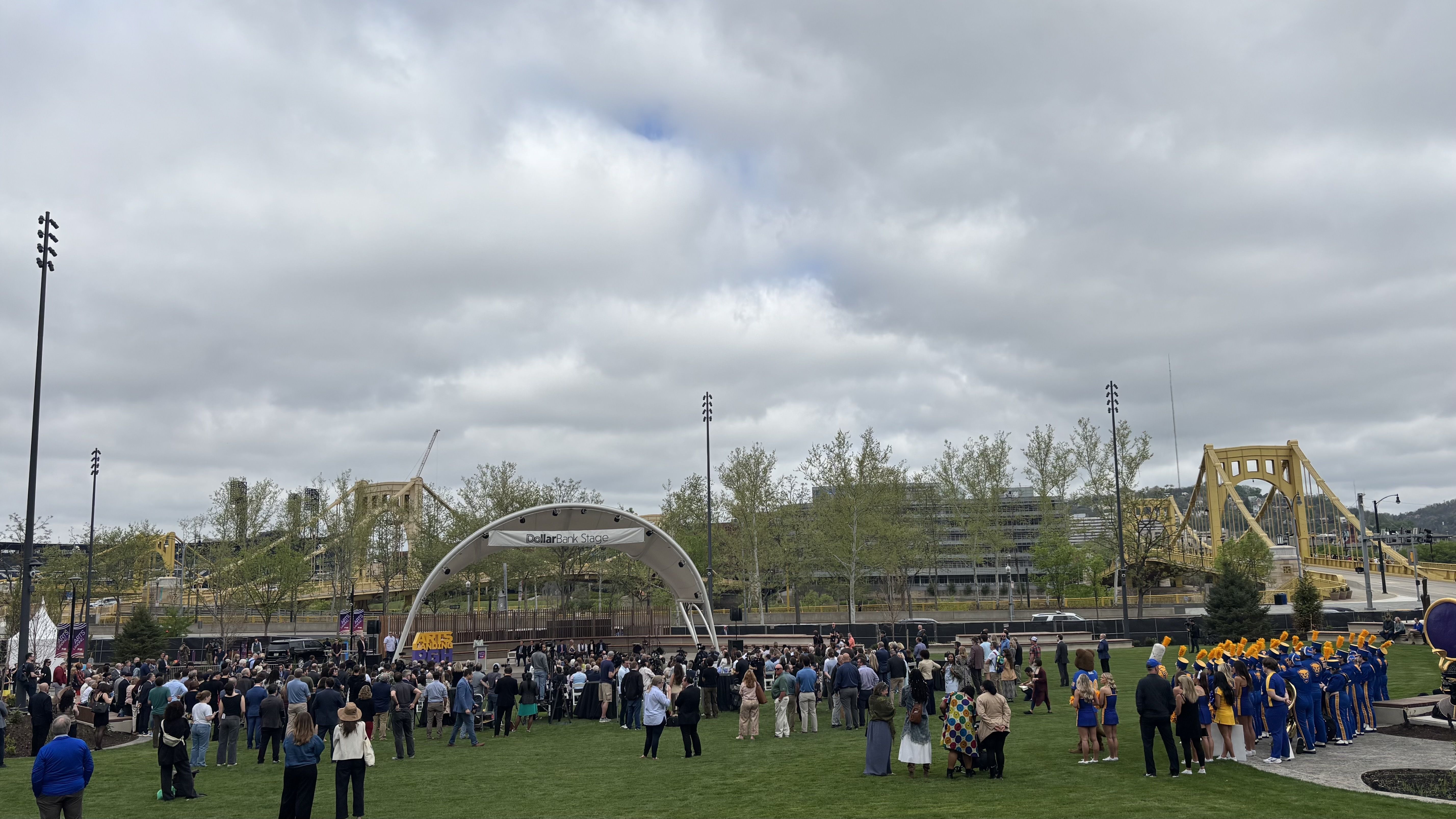 Overcast day on a grassy plaza; a crowd gathers around a white arched stage labeled "Dollar Bank Stage". To the right, a blue-and-yellow marching band stands with a large yellow bridge in the distance.