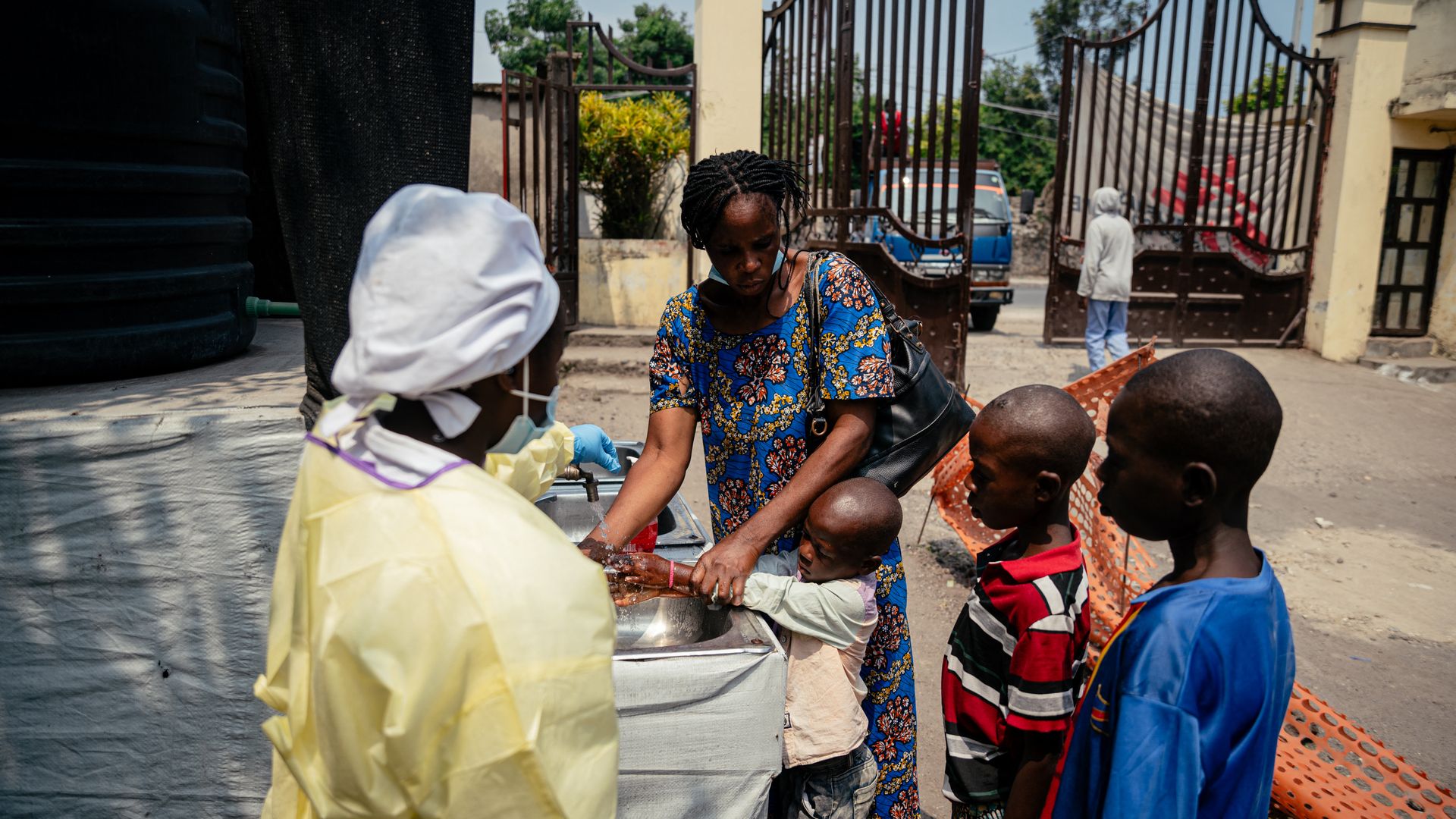 A woman wearing a colorful dress helps three young children wash their hands while a person wearing a mask and other protective equipment watches.