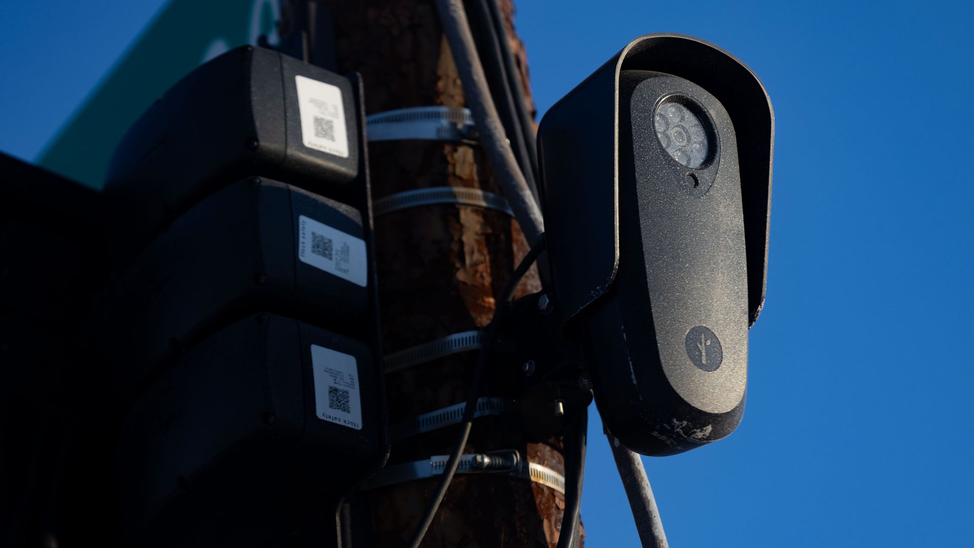Close-up of a street surveillance camera mounted on a rusty pole, housed in a black enclosure with a circular lens; cables and zip ties run along the pole against a blue sky.