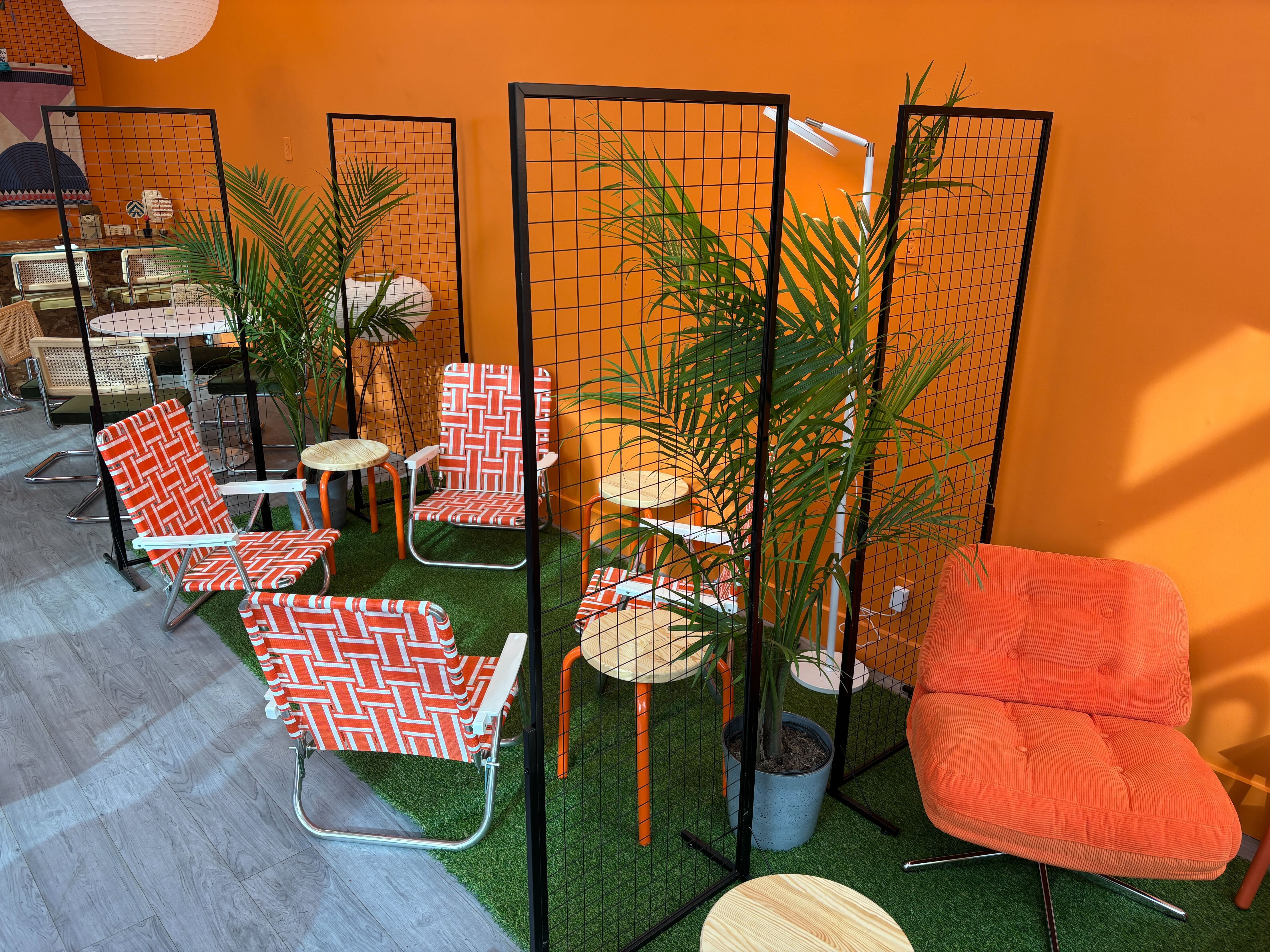 Indoor seating area with bright orange walls, orange and white patterned chairs, black metal grid room dividers, green plants, small round wooden tables, and an orange cushioned lounge chair.