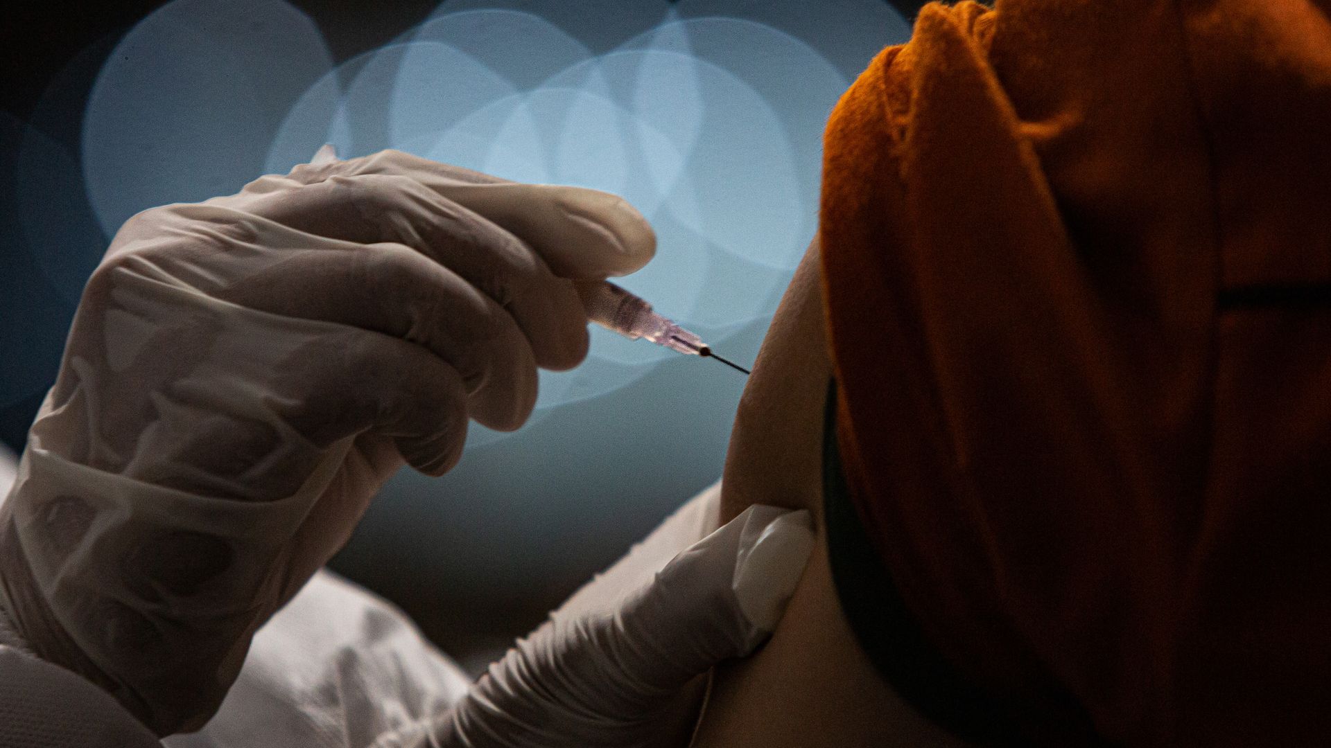 Photo of a gloved hand administering a vaccine shot to a person in an orange shirt