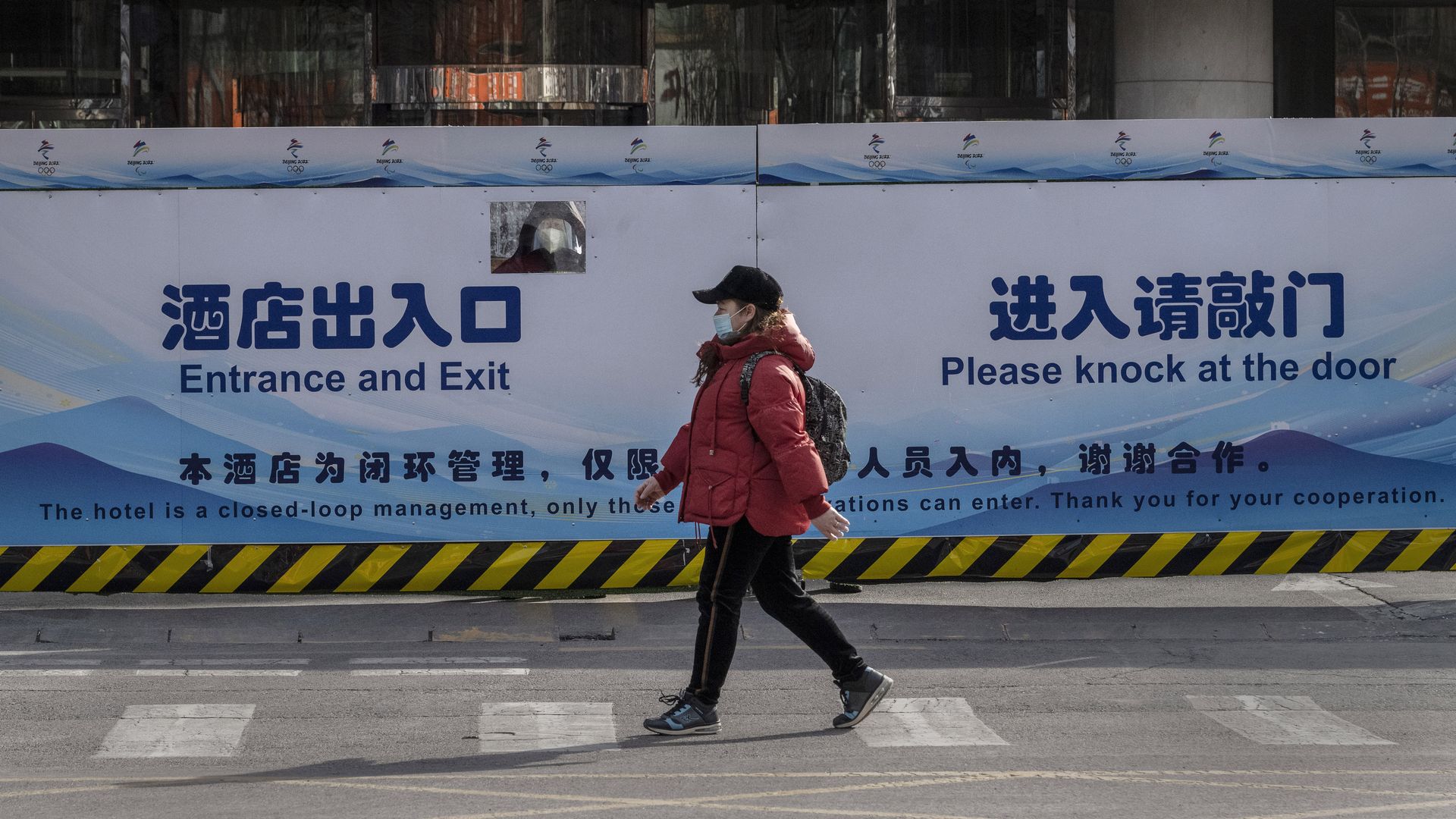 : A guard looks out through a small window from behind a gate as a pedestrian walks by in front of a fenced-in hotel being used as part of the "closed loop" system for the Beijing Winter Olympics.