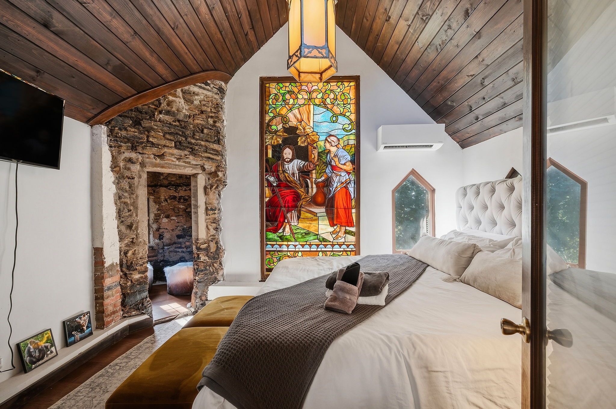 Loft-style bedroom with a dark wooden vaulted ceiling, exposed stone wall, and a tall stained-glass window centerpiece. A hanging lantern, white walls, and a bed with white and gray bedding.