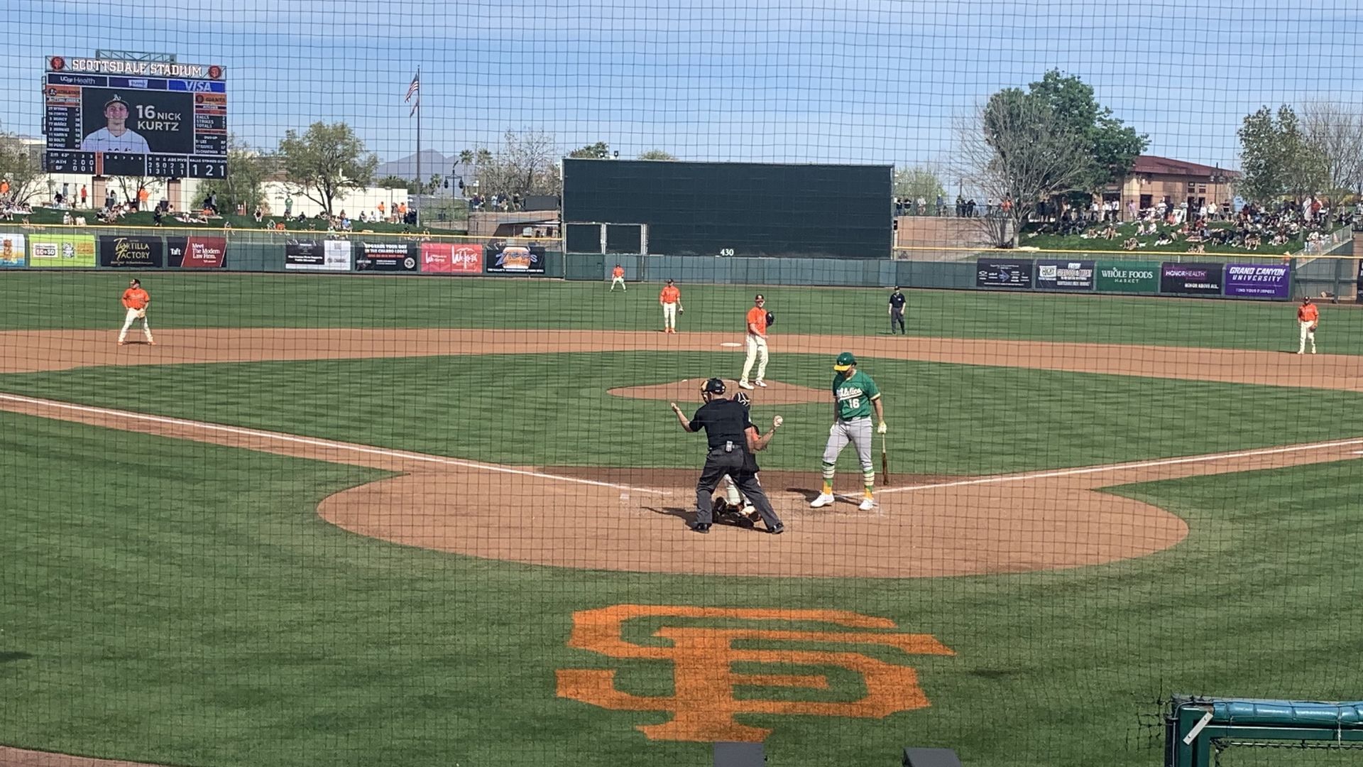Baseball game at Scottsdale Stadium under clear blue sky, Oakland Athletics batter in green, Giants players in orange on field, scoreboard showing player Nick Kurtz #16, crowd in stands.