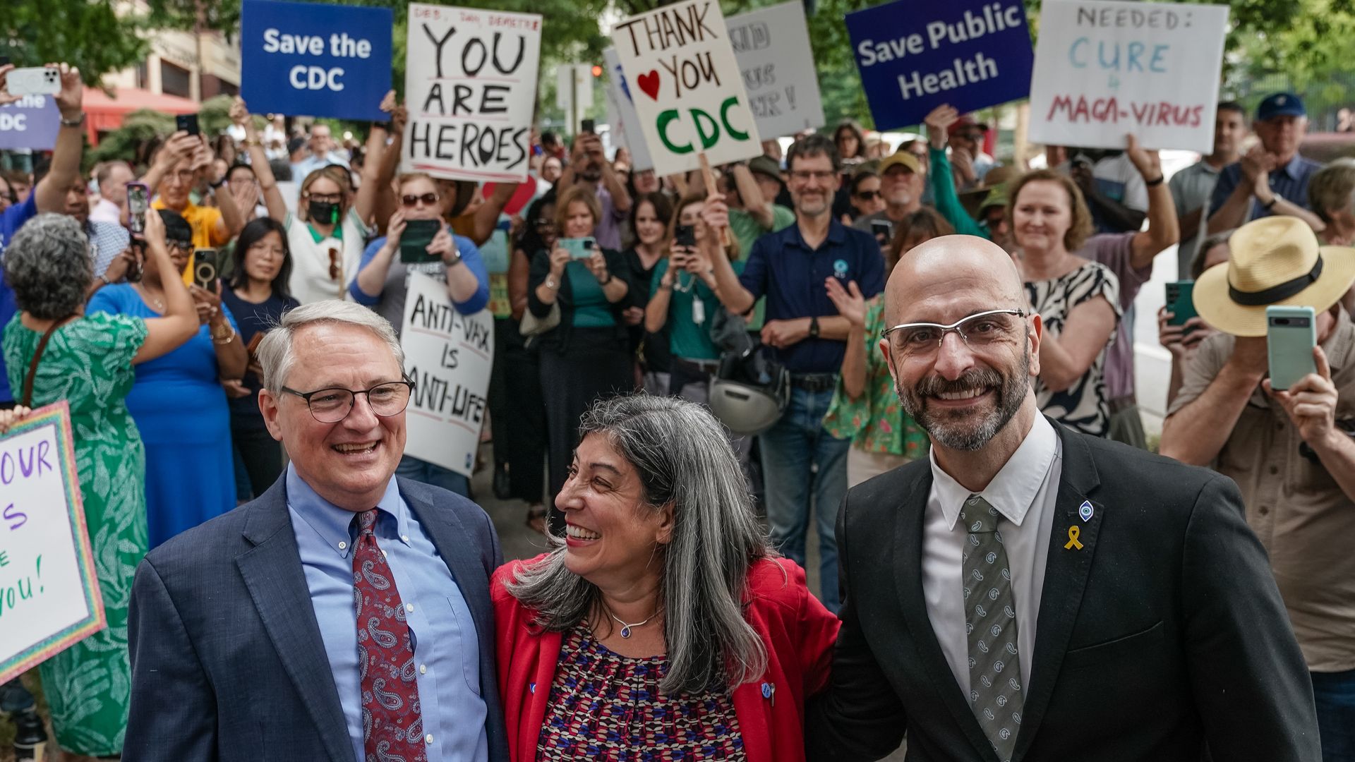 Crowd at rally holding signs like "Save the CDC," "You Are Heroes," and "Thank You CDC," with three smiling adults in front, two men in suits and a woman in a red jacket, outdoors with green trees.