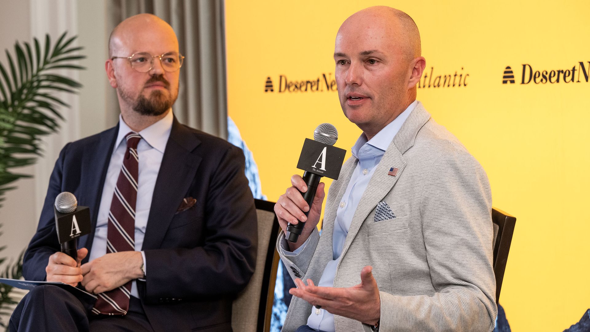Two men in suits sit on a panel; the right speaks into a microphone, the left listens. Bright yellow backdrop with logos and a plant visible on the left.
