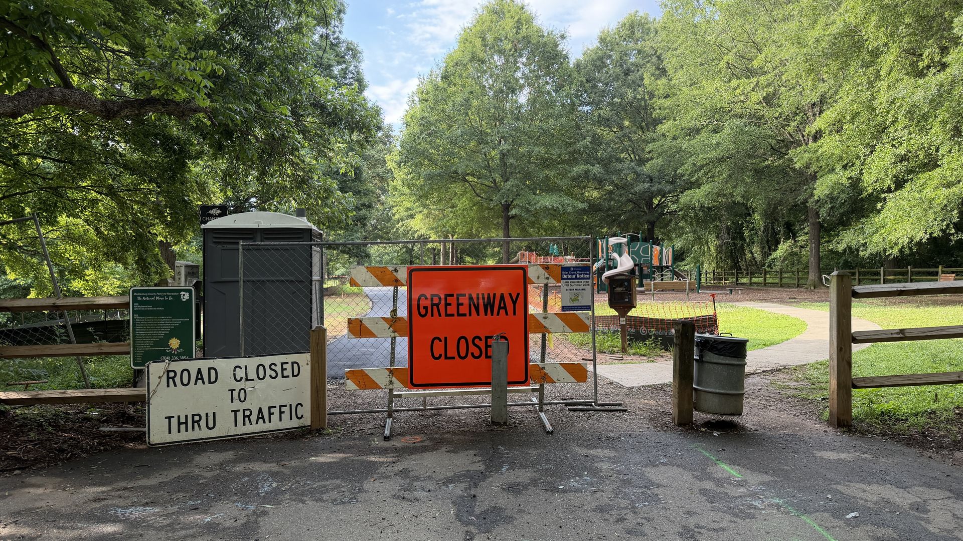 Sunlit park scene with barriers and an orange "Greenway Closed" sign blocking a path; a chain-link fence, portable toilet, and road-closure signs, with trees and a playground in the distance.
