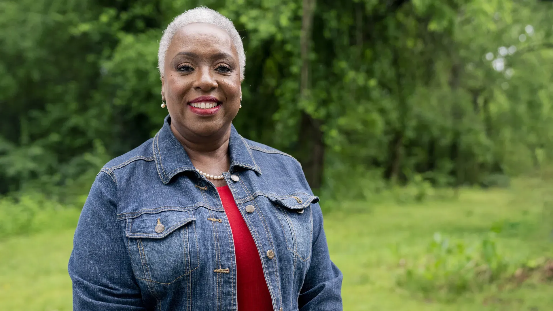 Smiling woman with short gray hair outdoors in a green, wooded area. She wears a denim jacket over a red top, with a pearl necklace and matching pearl earrings.