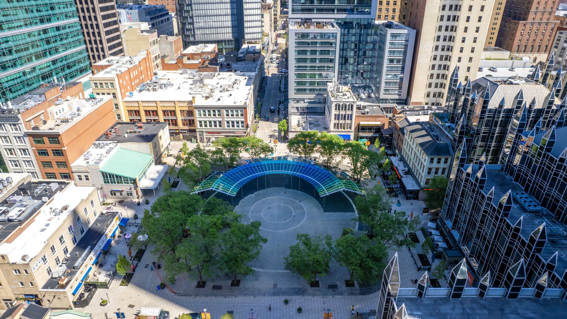 Aerial view of a downtown block with glass and brick towers surrounding a circular plaza. A blue curved glass canopy arches over the space, with green trees along the edges.