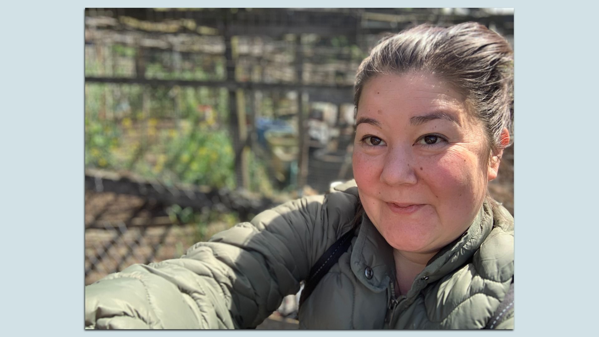 Jill Wasburg looks at the camera while standing in front of garden plots, wearing an olive green puffer coat.