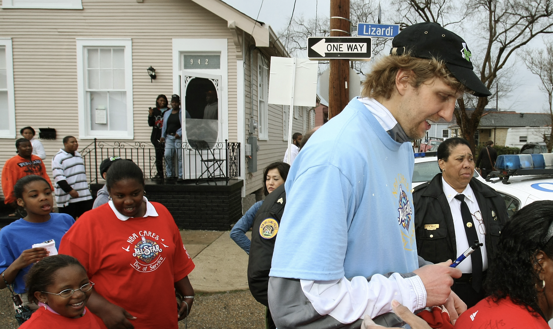 Man in light blue NBA Cares t-shirt and black cap signing autographs for a group of girls wearing red and blue shirts, on a street corner with a beige house and police officer nearby under a one-way sign.