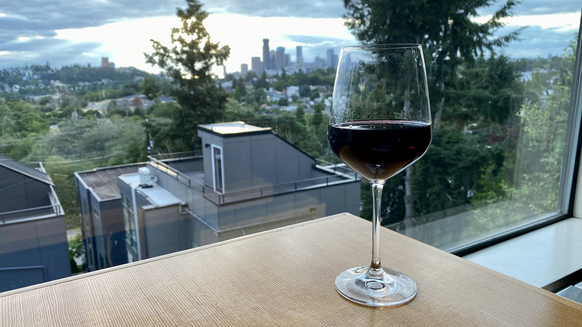A glass of red wine on a wooden bar next to a window, with views of trees and the city in the distance.