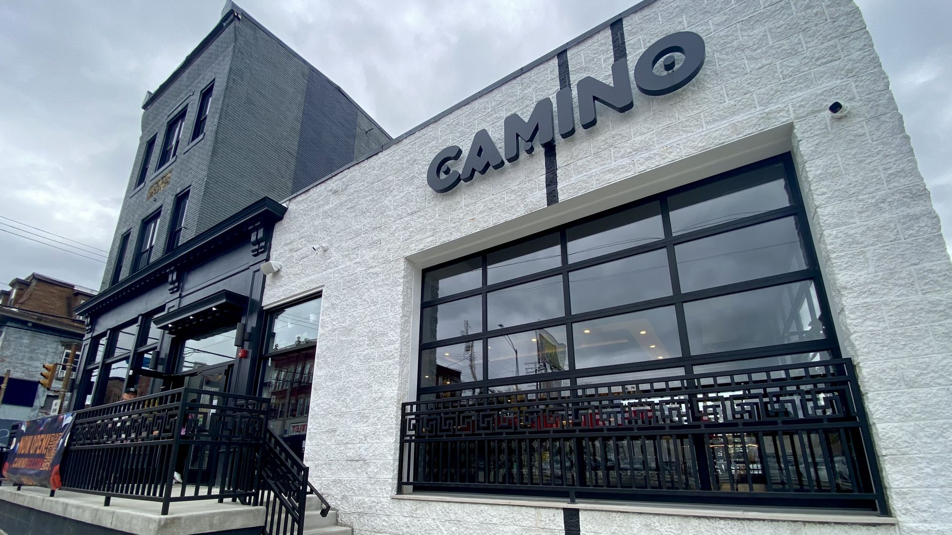 Exterior of a white brick building with large windows and black railing, featuring the sign "CAMINO" above the window, under an overcast sky.